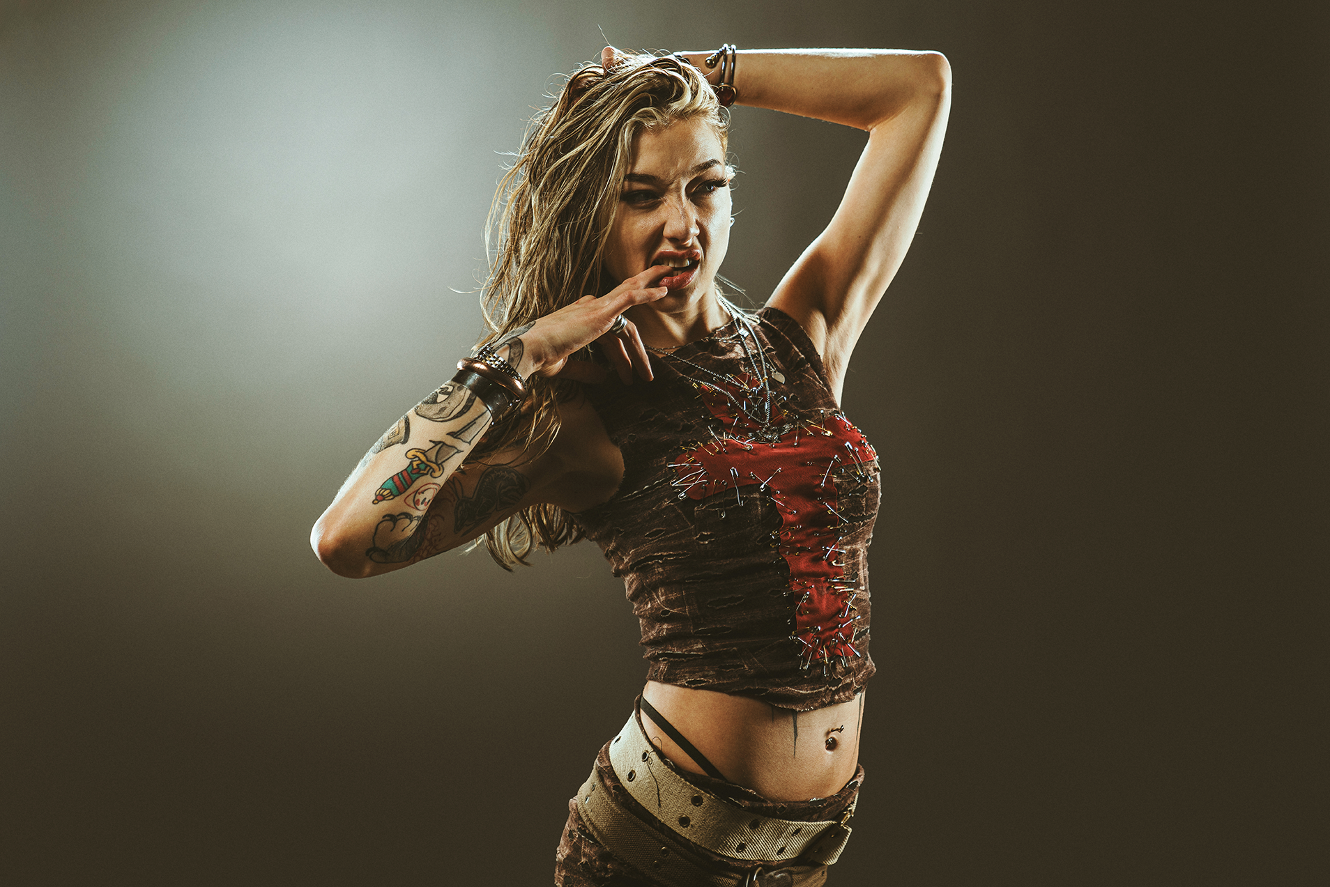 Young woman with tattoos posing confidently in a studio with a dark background, wearing layered jewelry, a distressed crop top with a red heart design, and patterned pants.