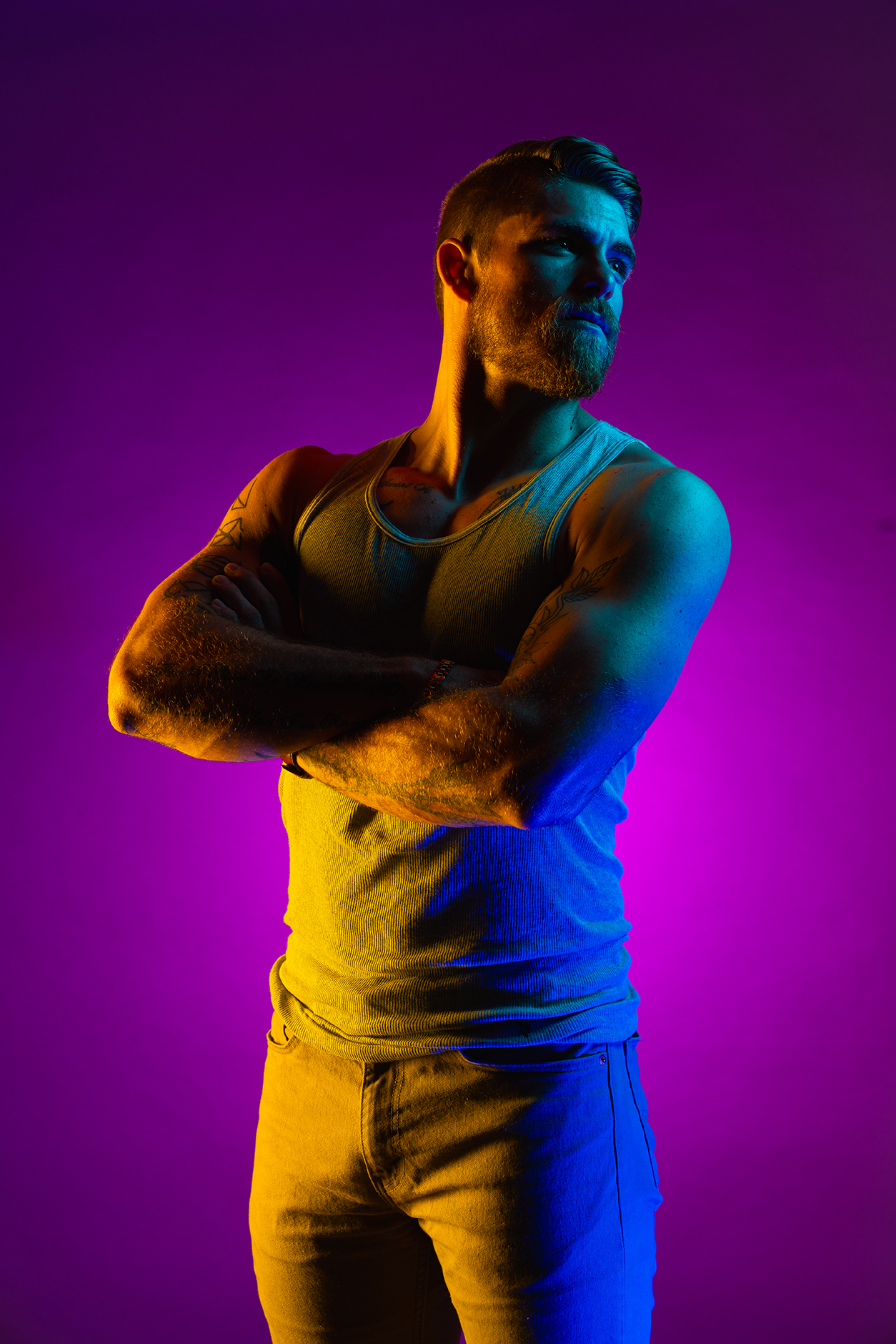 A bearded man with tattoos on his arms wearing a gray tank top and pants, standing with arms crossed against a purple and blue gradient background.