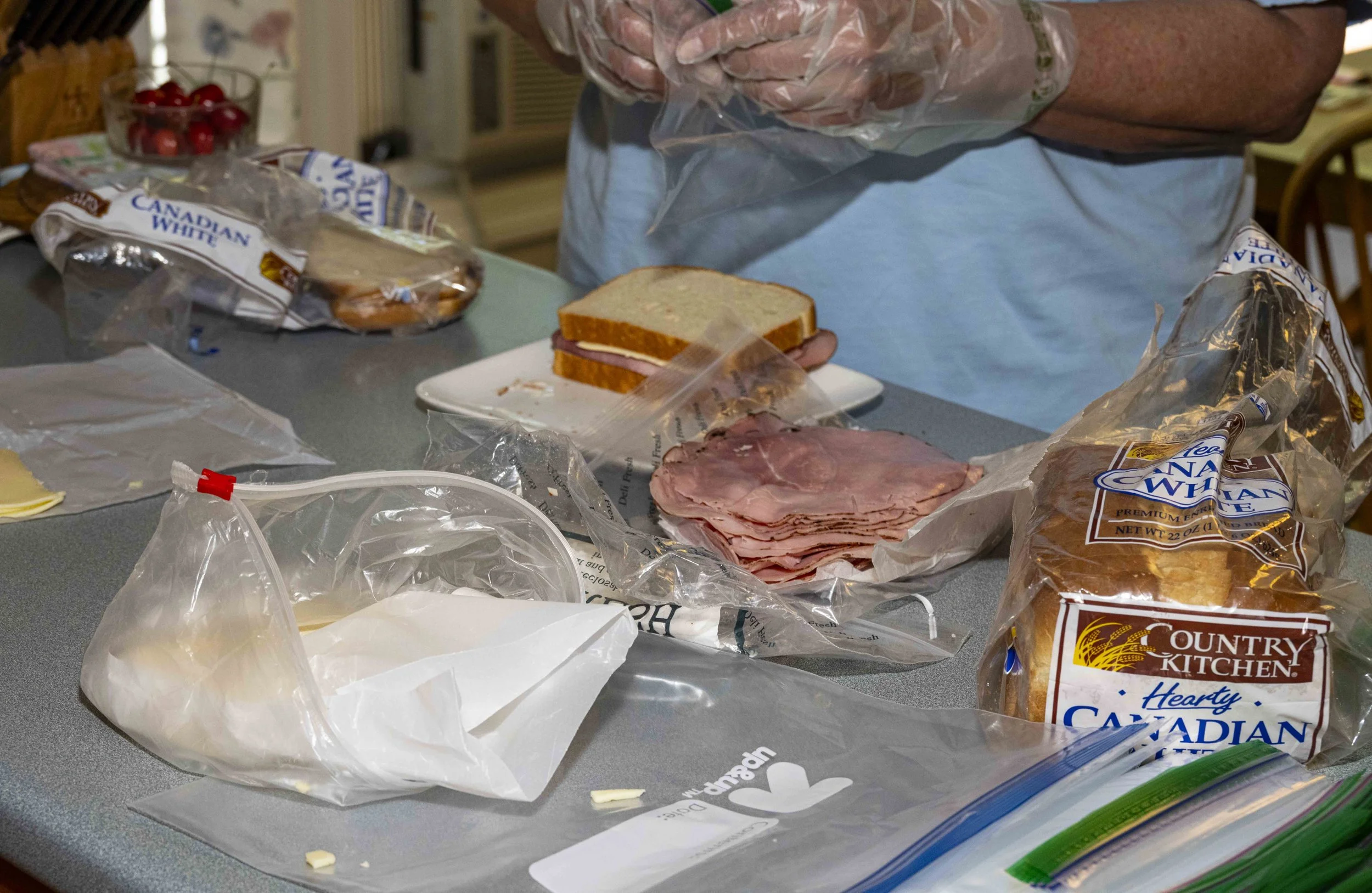Person preparing sandwiches with bread, ham, and cheese on a kitchen counter, with various sliced bread packages and cherry tomatoes visible.