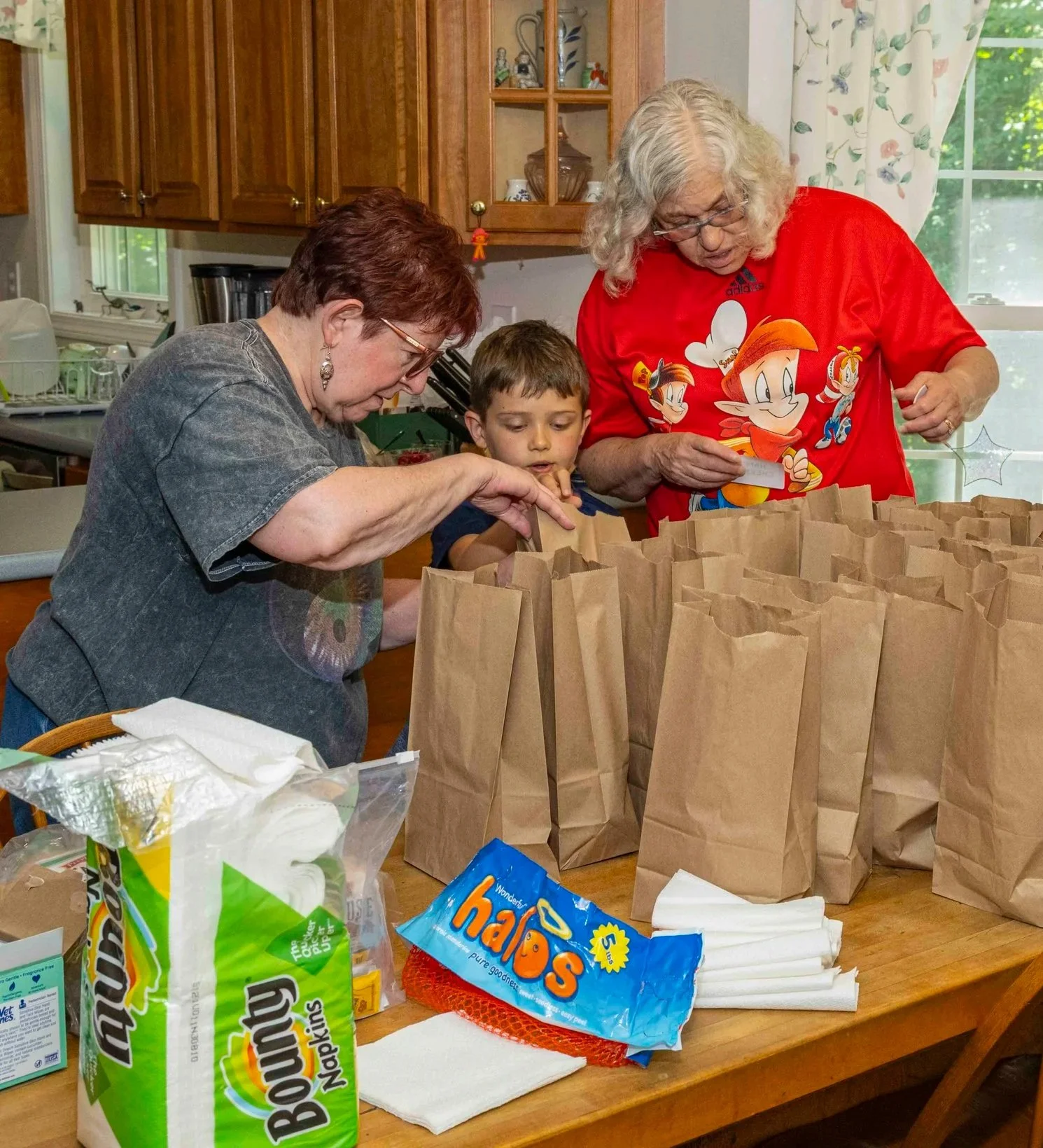 Three people, two women and a young boy, packing brown paper bags on a kitchen table, with supplies like napkins and bags of marshmallows around.