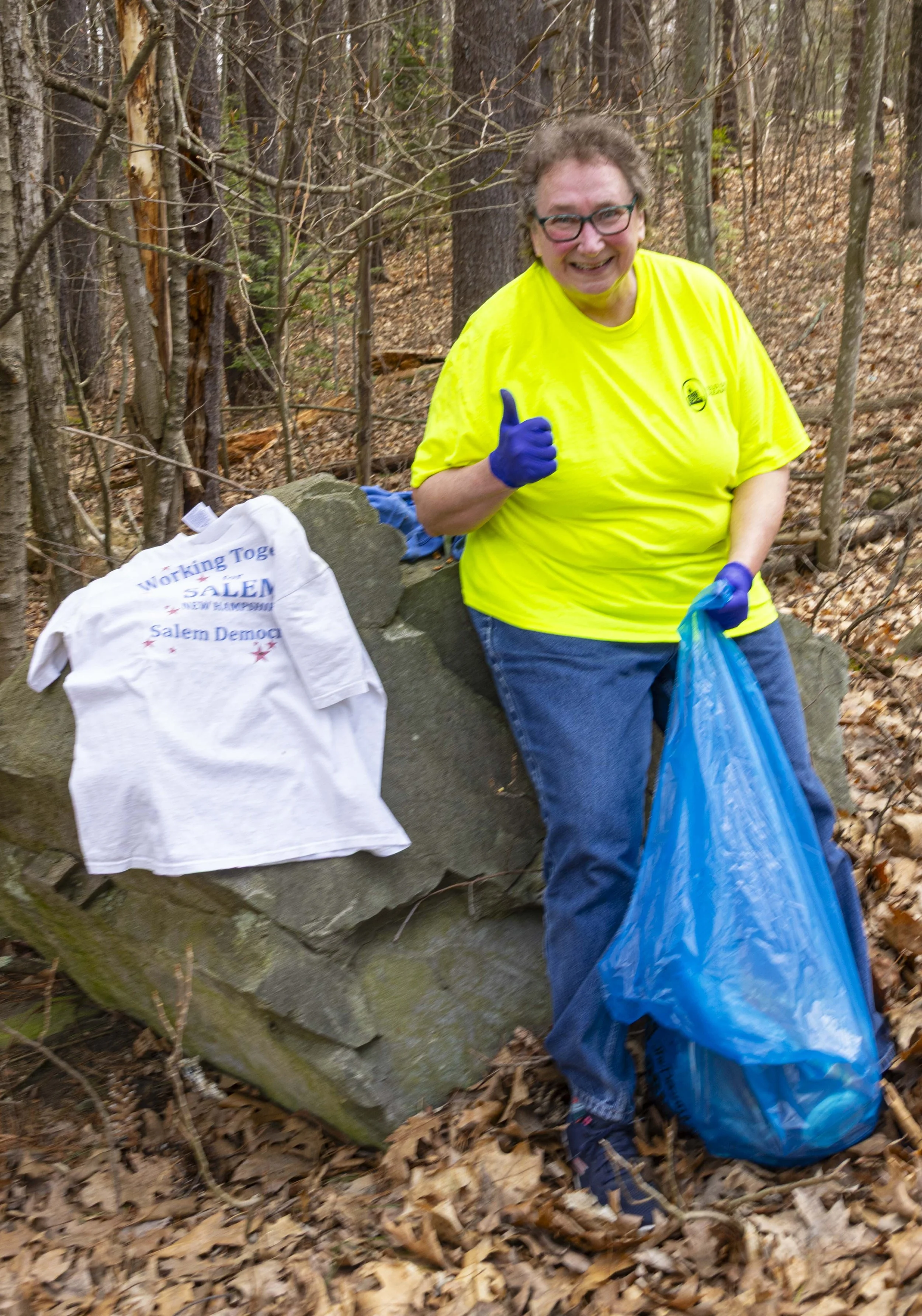 Woman in a yellow shirt and blue gloves giving a thumbs-up while collecting trash in a forest, with a blue trash bag and white t-shirt on a rock.