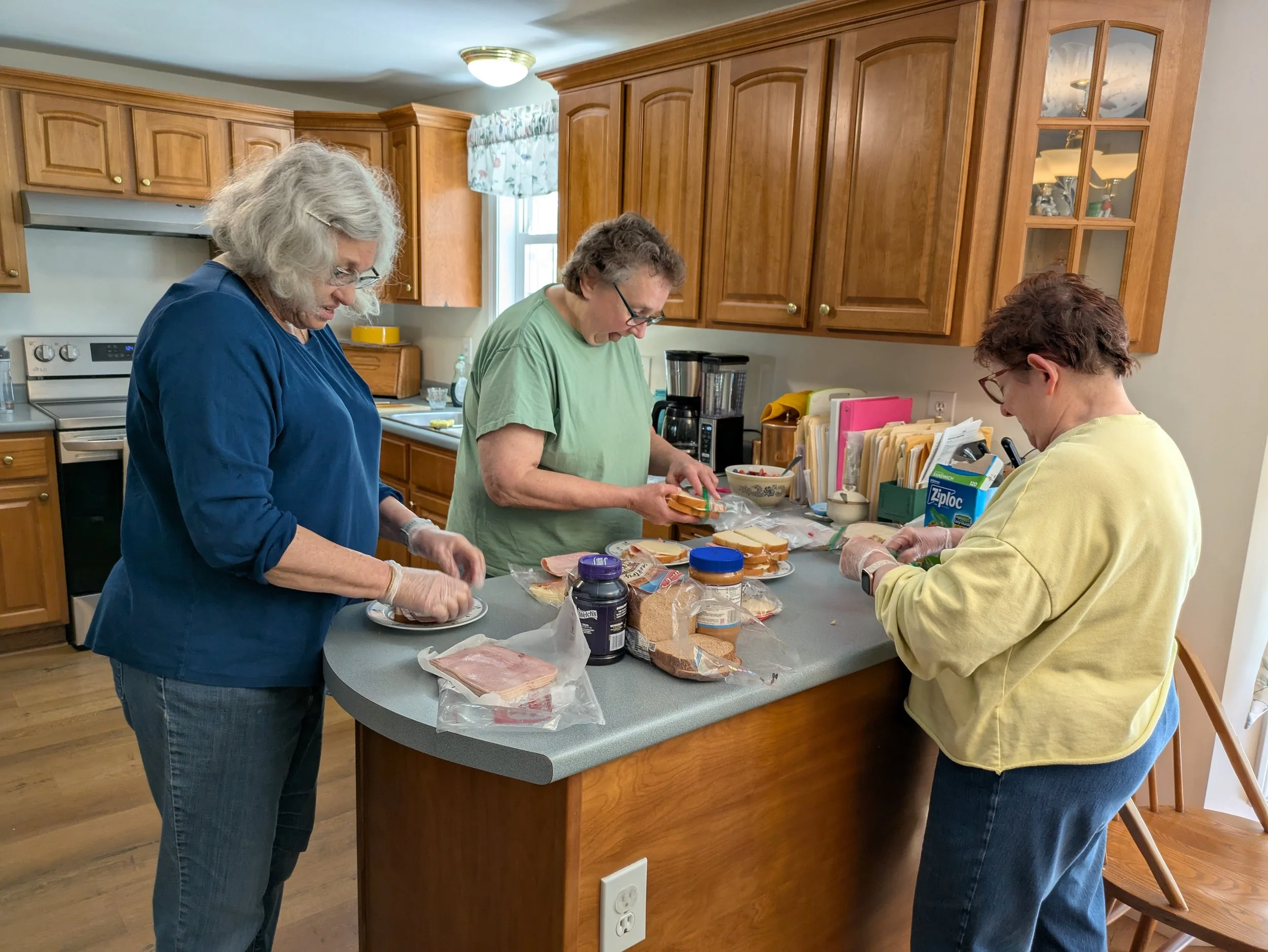 Three women preparing sandwiches in a kitchen, with various sandwich ingredients on the countertop.