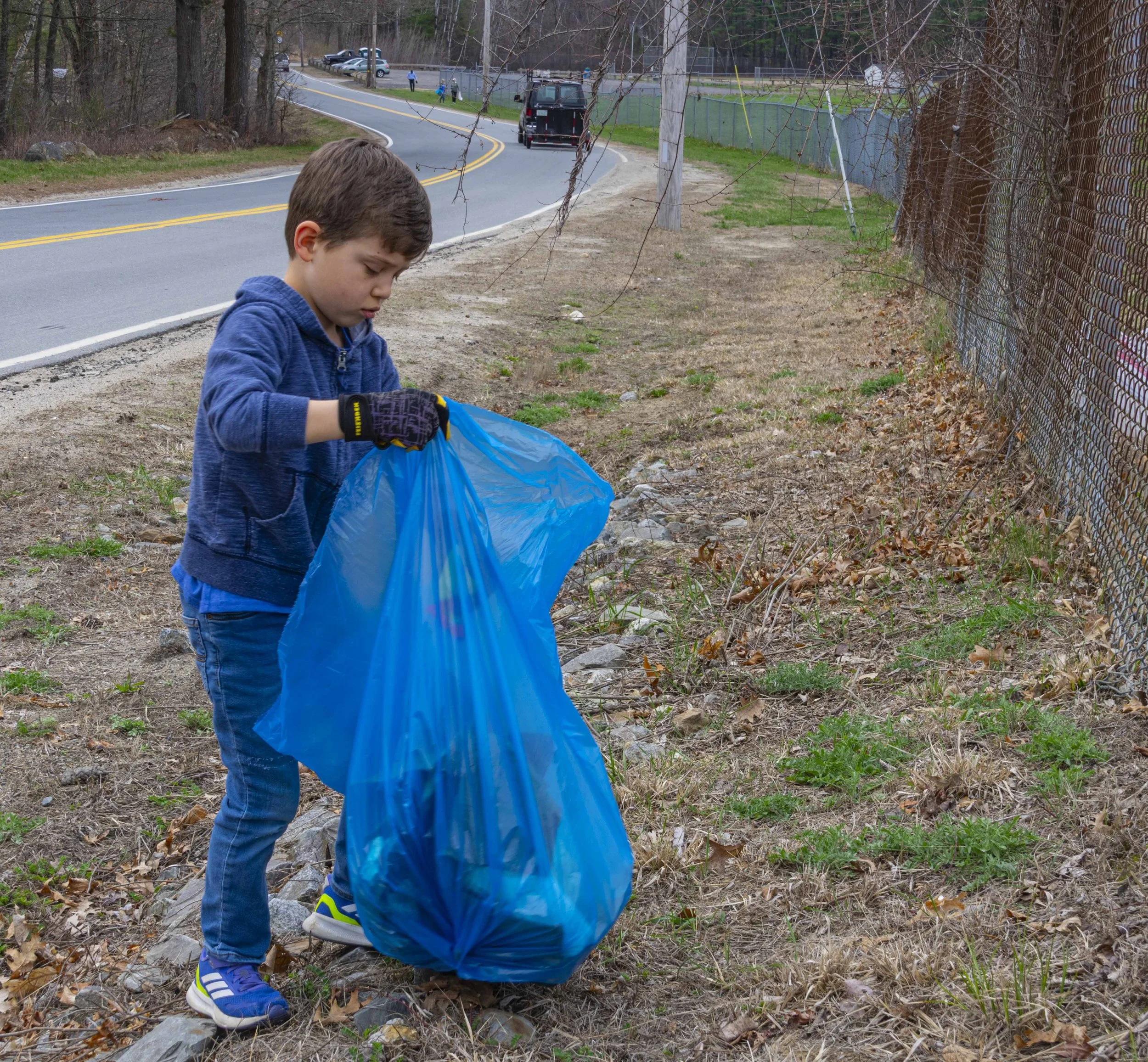 Young boy in a blue hoodie and jeans picks up trash with a blue garbage bag along a roadside.
