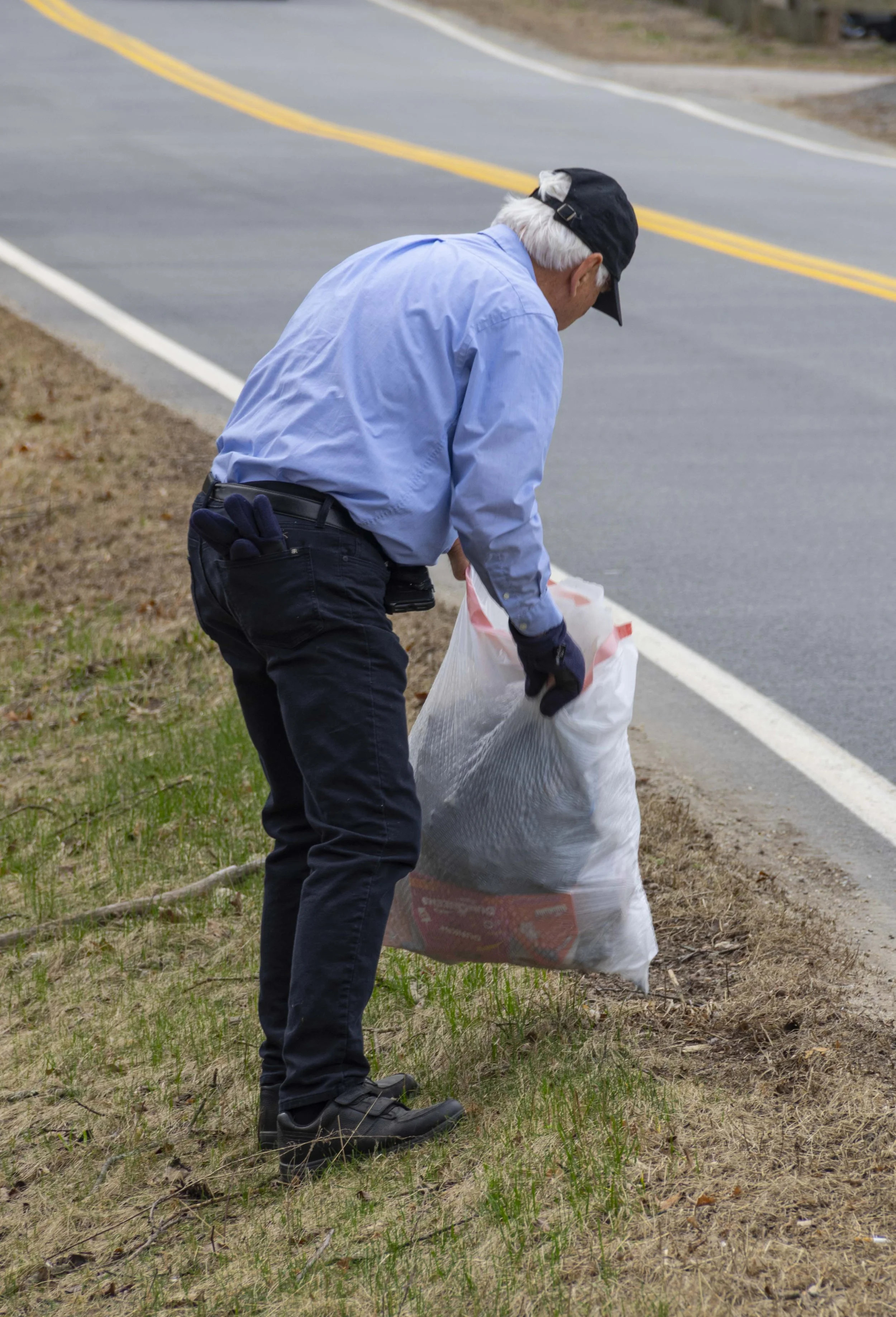 An elderly man wearing a light blue shirt, black pants, and a black cap picks up trash from the roadside into a plastic bag.