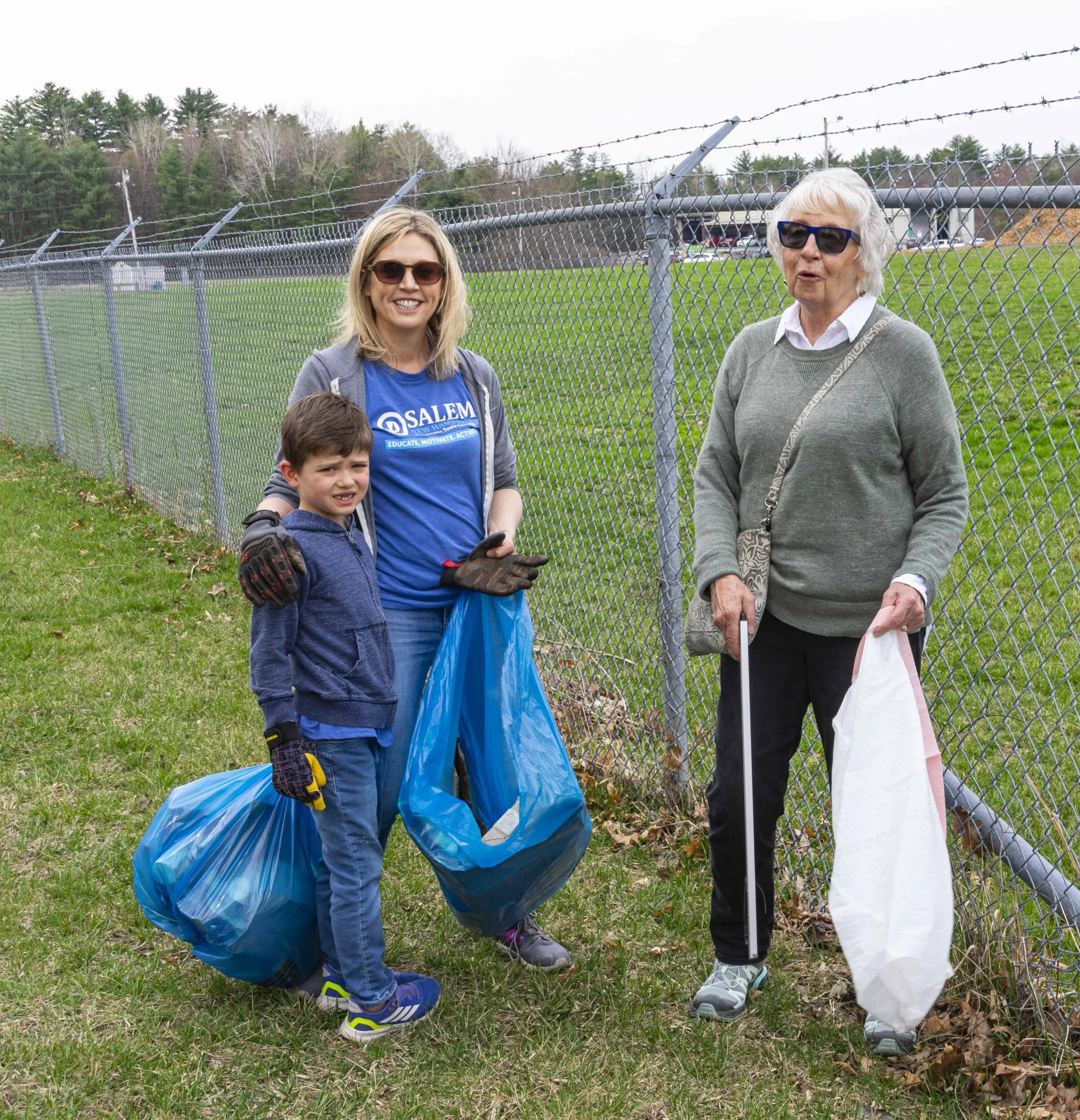 Three people standing outdoors by a chain-link fence, participating in a community cleanup. One woman with blonde hair wearing sunglasses, a blue "Salem" shirt, and gloves, holds trash bags. A young boy with dark hair, a blue hoodie, and gloves stand