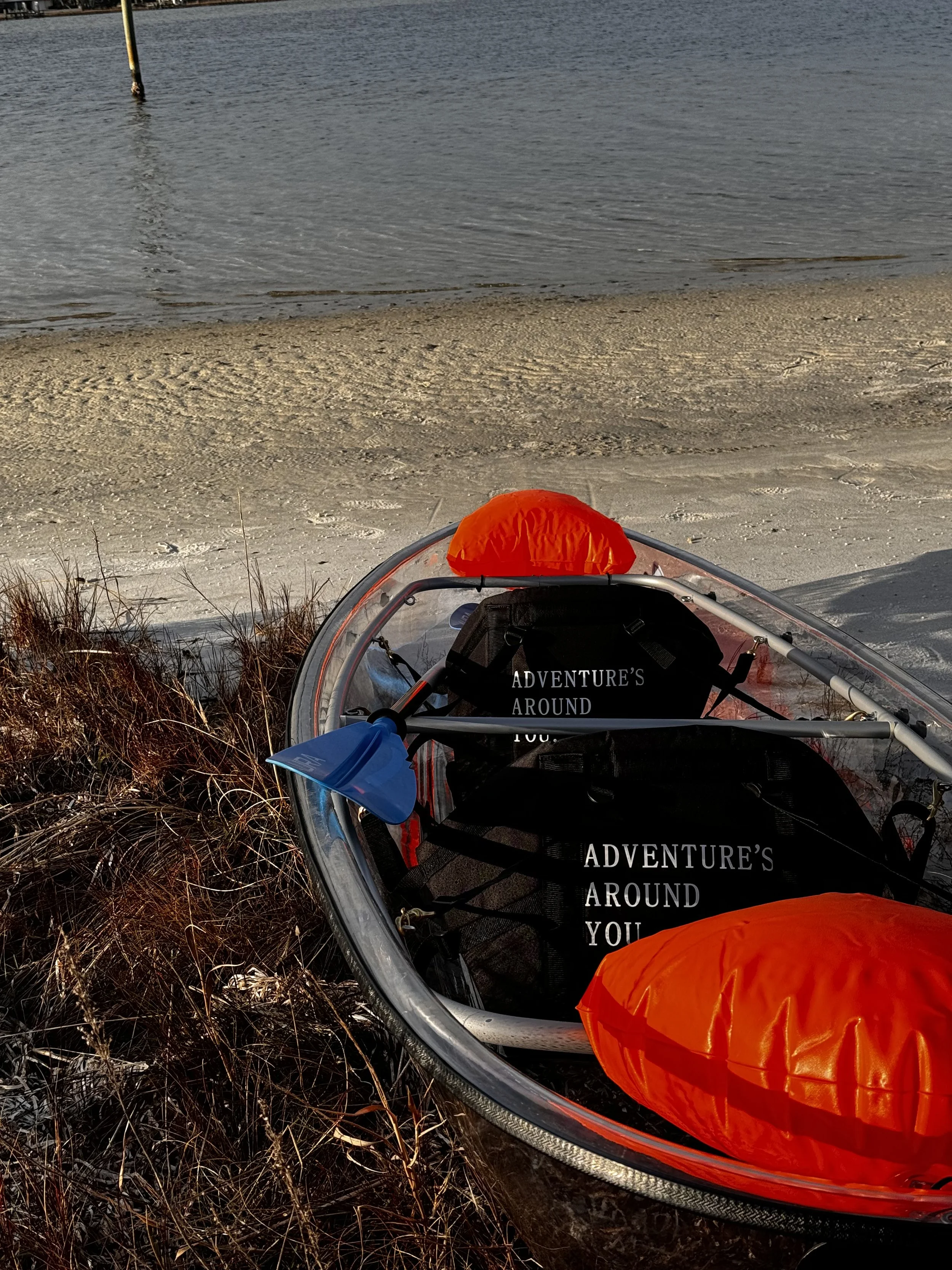 An inflatable boat on a lakeshore with a life vest, paddle, and a black bag that says 'Adventure's Around You'. The lake's water and sandy shore are visible in the background.