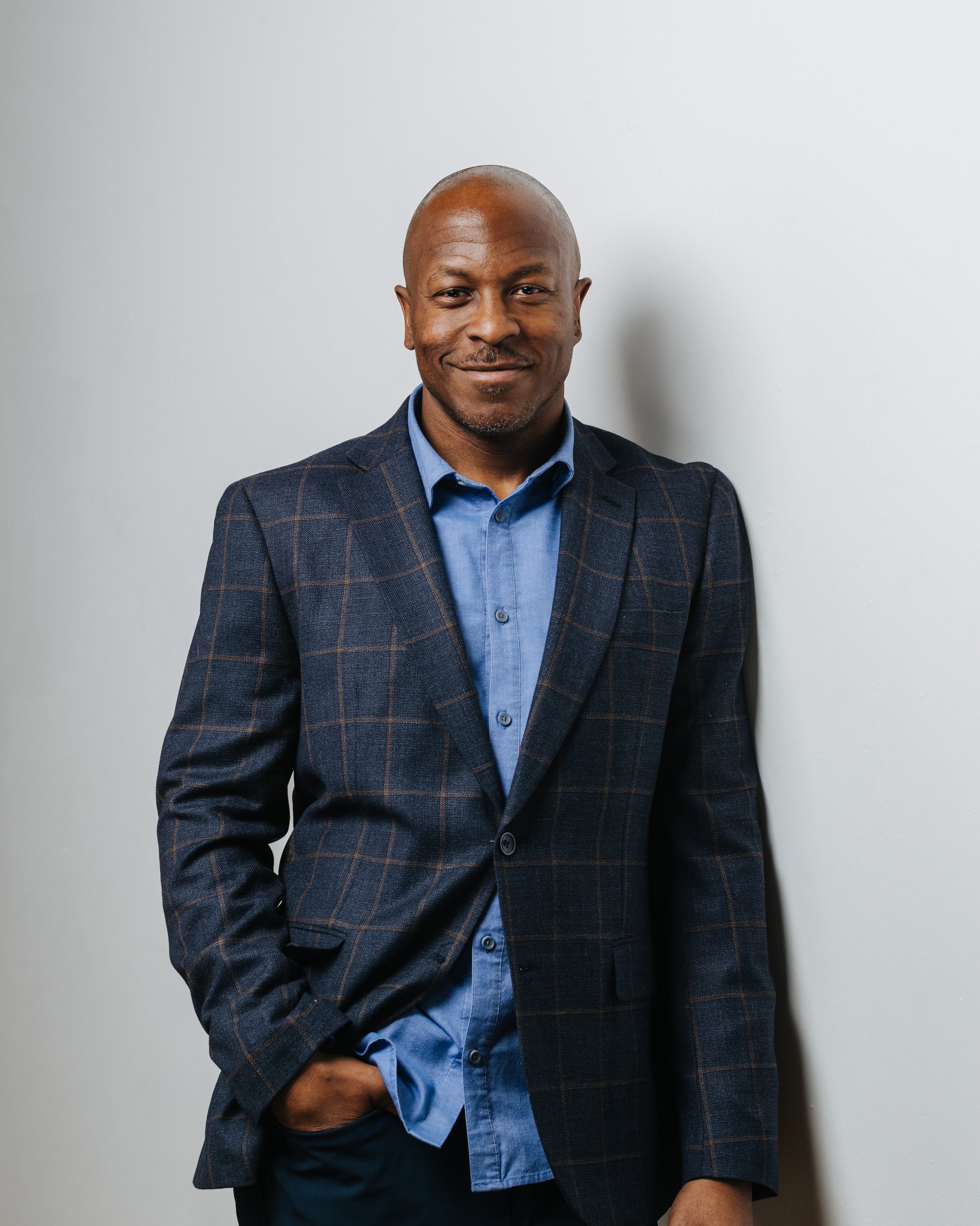A man in a blue shirt and checked blazer standing against a white wall, with one hand in his pocket and a slight smile.