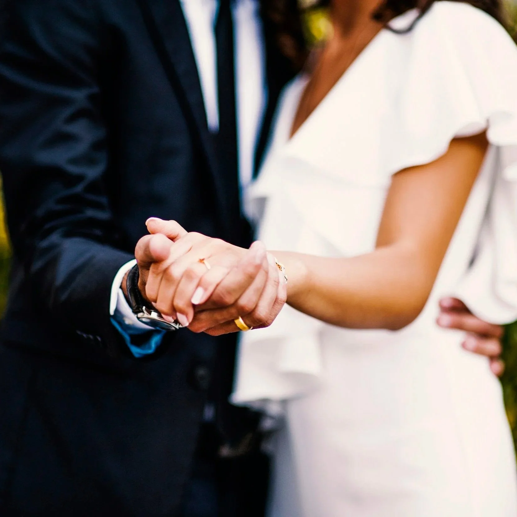 Close-up of hands of a bride and groom holding each other's hands during a wedding, wearing wedding rings and dressed in formal attire.