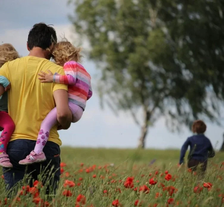 A man wearing a yellow shirt carrying a young girl in colorful striped clothing through a field of red flowers with a tree and another child in the background.