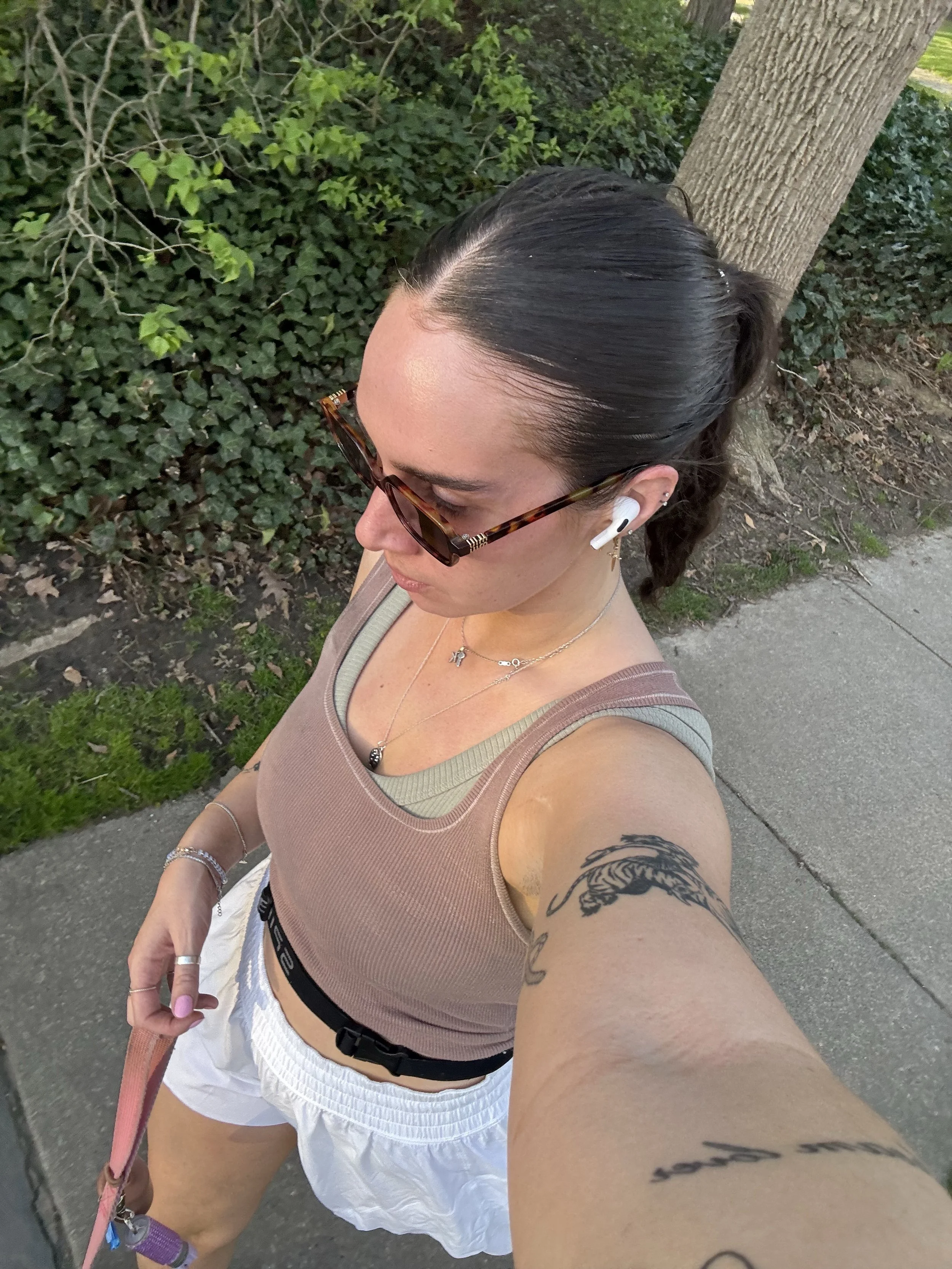 A young woman taking a selfie outdoors on a sidewalk next to a tree and green shrubbery. She has dark hair tied back, is wearing sunglasses, a beige tank top, white shorts, and wireless earbuds. She has tattoos on her left arm and wears jewelry, including necklaces and bracelets.