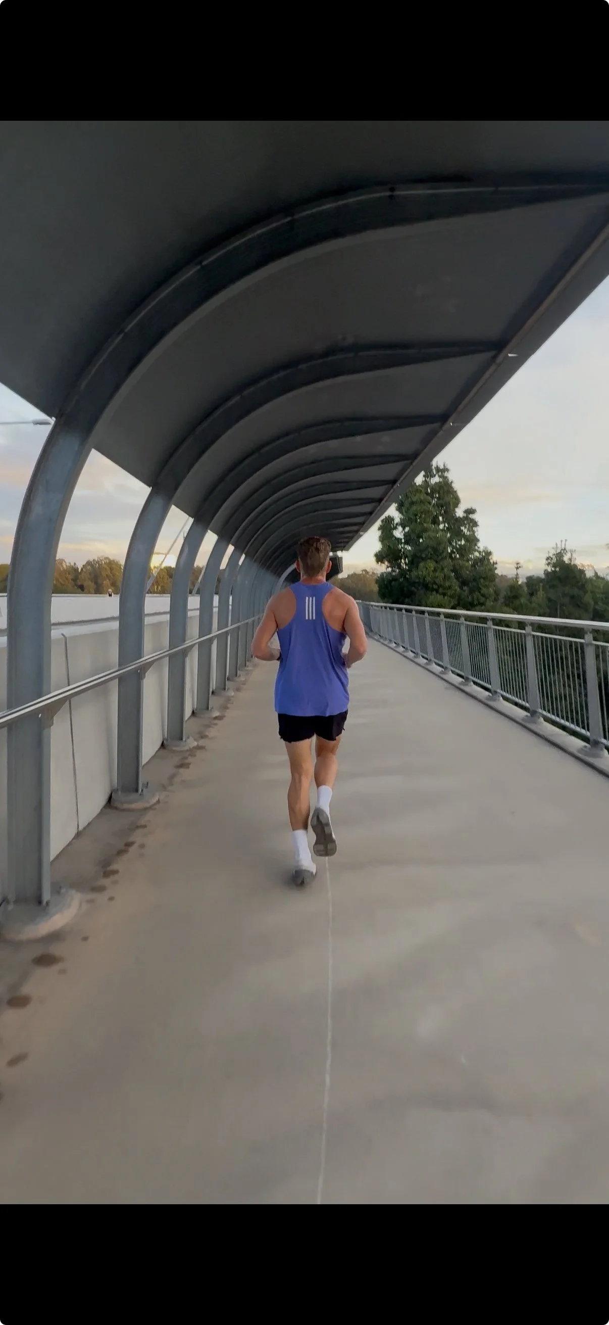 Person running on a covered pedestrian bridge during sunset, wearing a blue sleeveless shirt and black shorts.