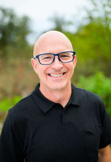 A smiling, bald man wearing glasses and a black polo shirt outdoors with greenery in the background.