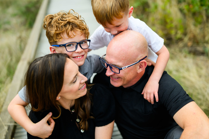 A family of four, including two children and their parents, smiling and playing together outdoors on a wooden walkway surrounded by greenery.