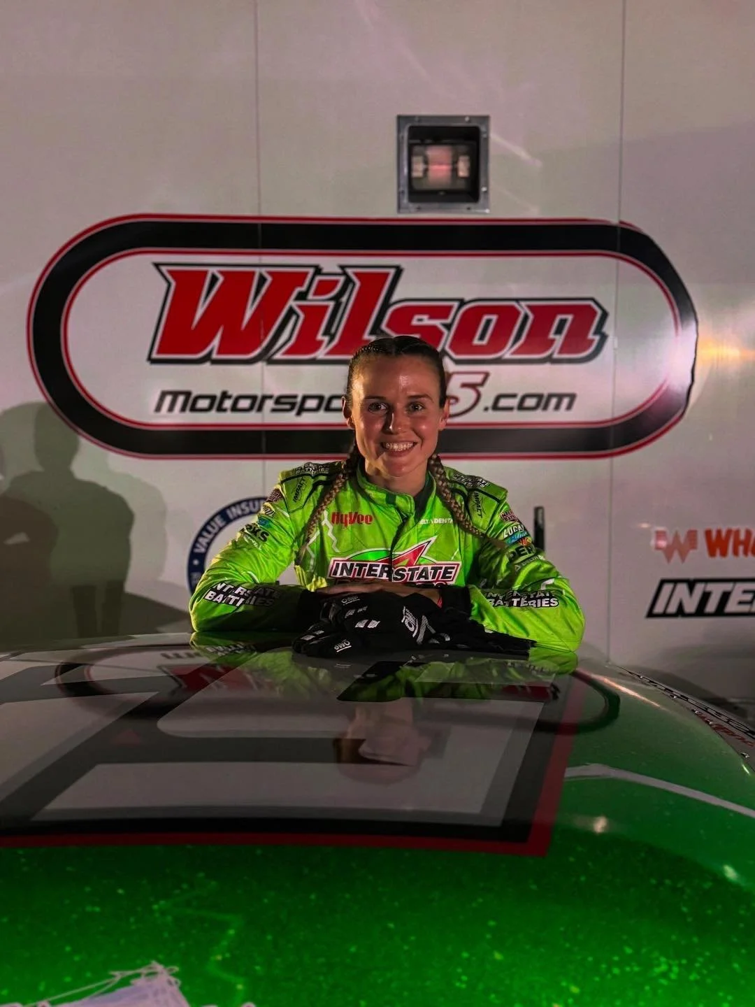 McKenna Haase Scott in a bright green racing suit sitting at a racing car with hands on the hood. Behind her is a large Wilson Motorsports.com sign.