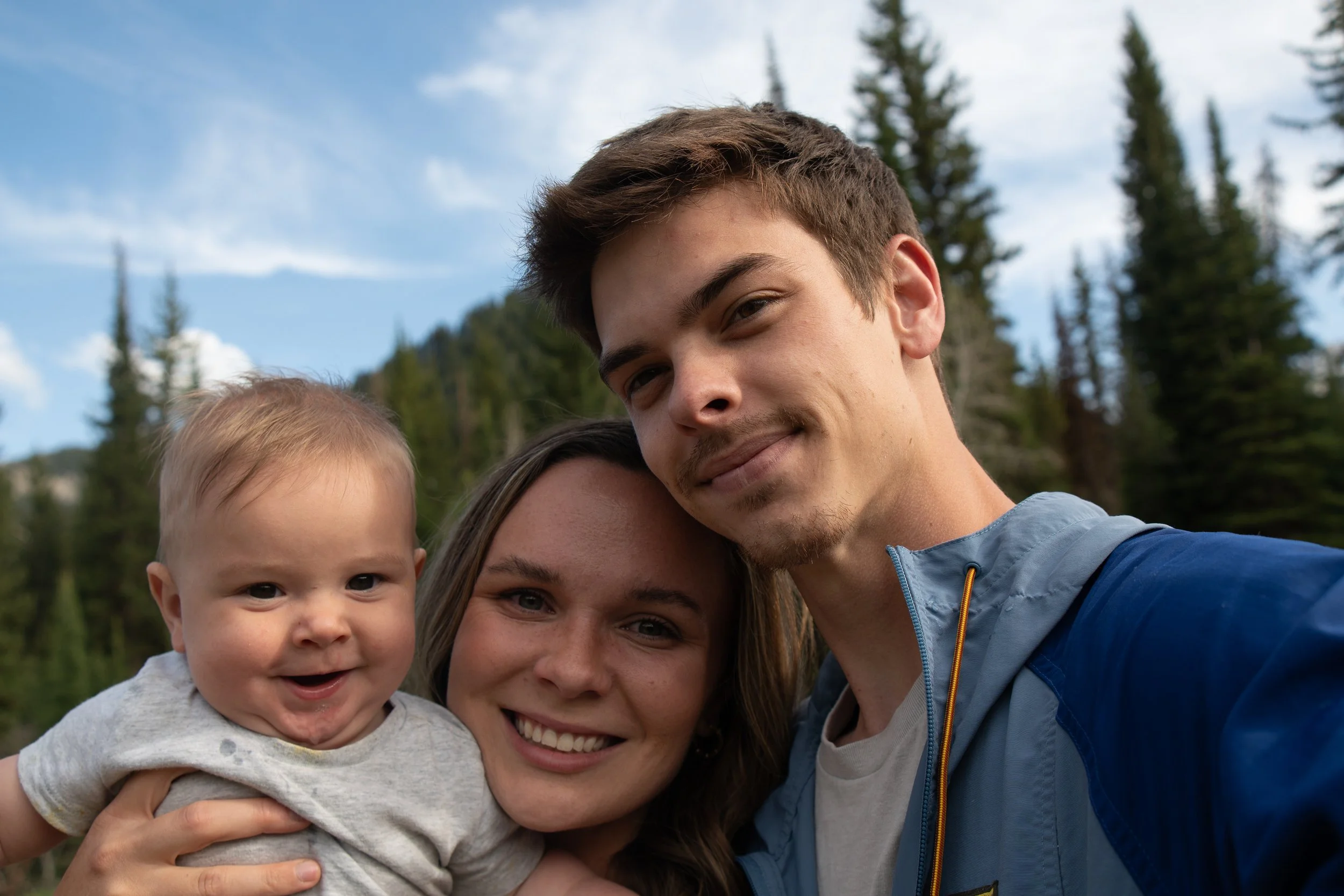 A smiling family of three taking a selfie outdoors in a forested area with tall pine trees and a cloudy sky.