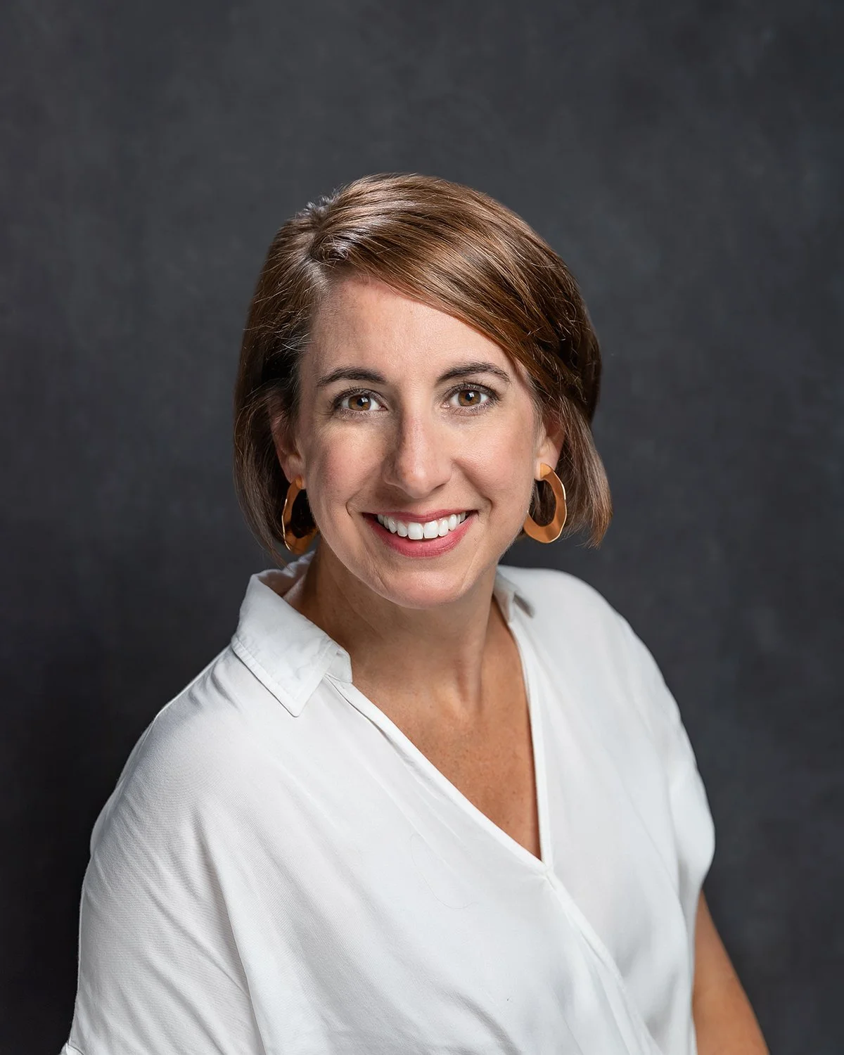 Close-up portrait of a woman with short brown hair, wearing a white blouse and orange hoop earrings, smiling against a dark gray background.