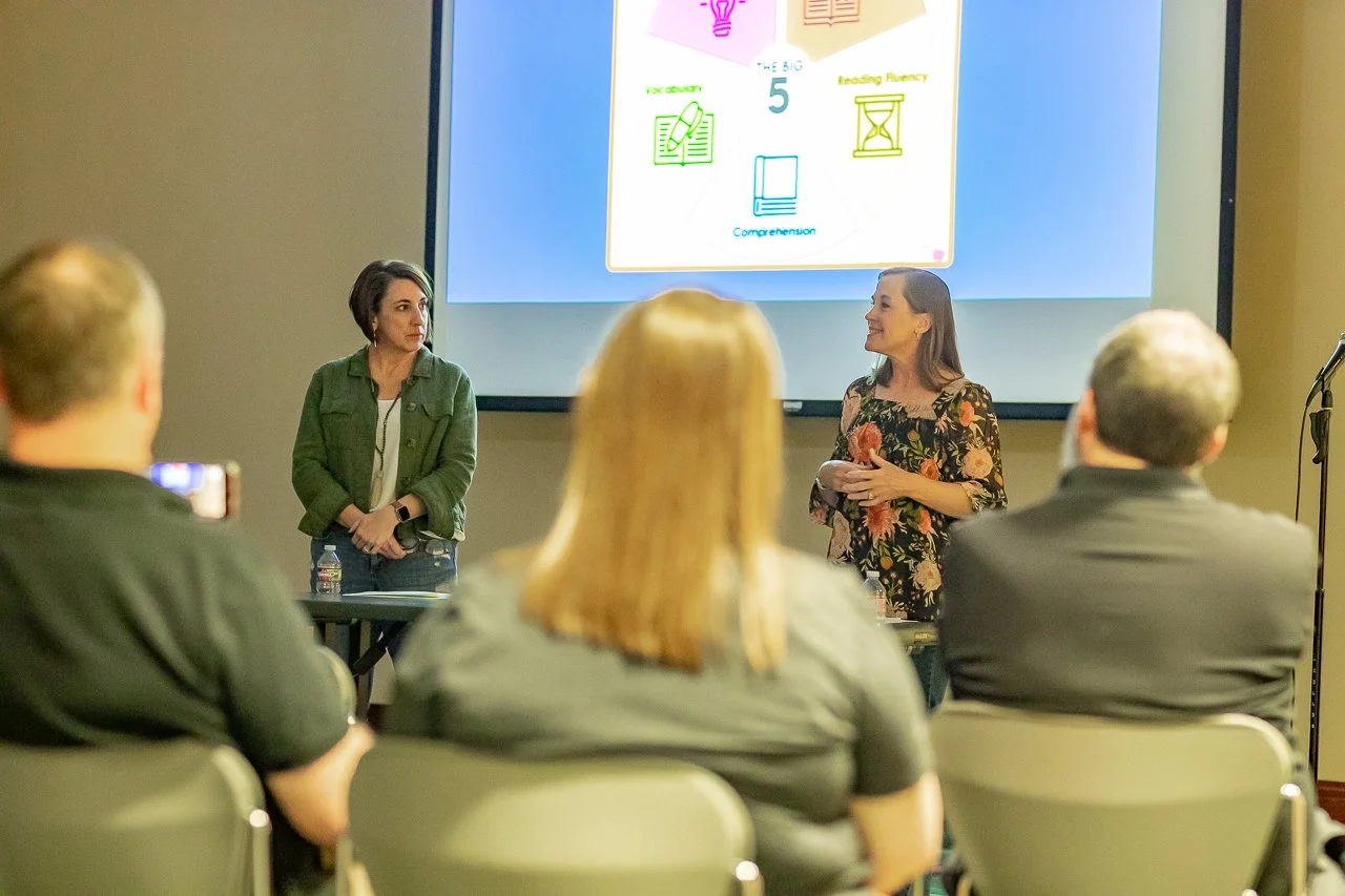Two women speaking at a presentation in a room with an audience. One woman is standing with her hands clasped, wearing a green jacket, and the other woman is gesturing with her hands, wearing a floral top. A large screen behind them displays simplist