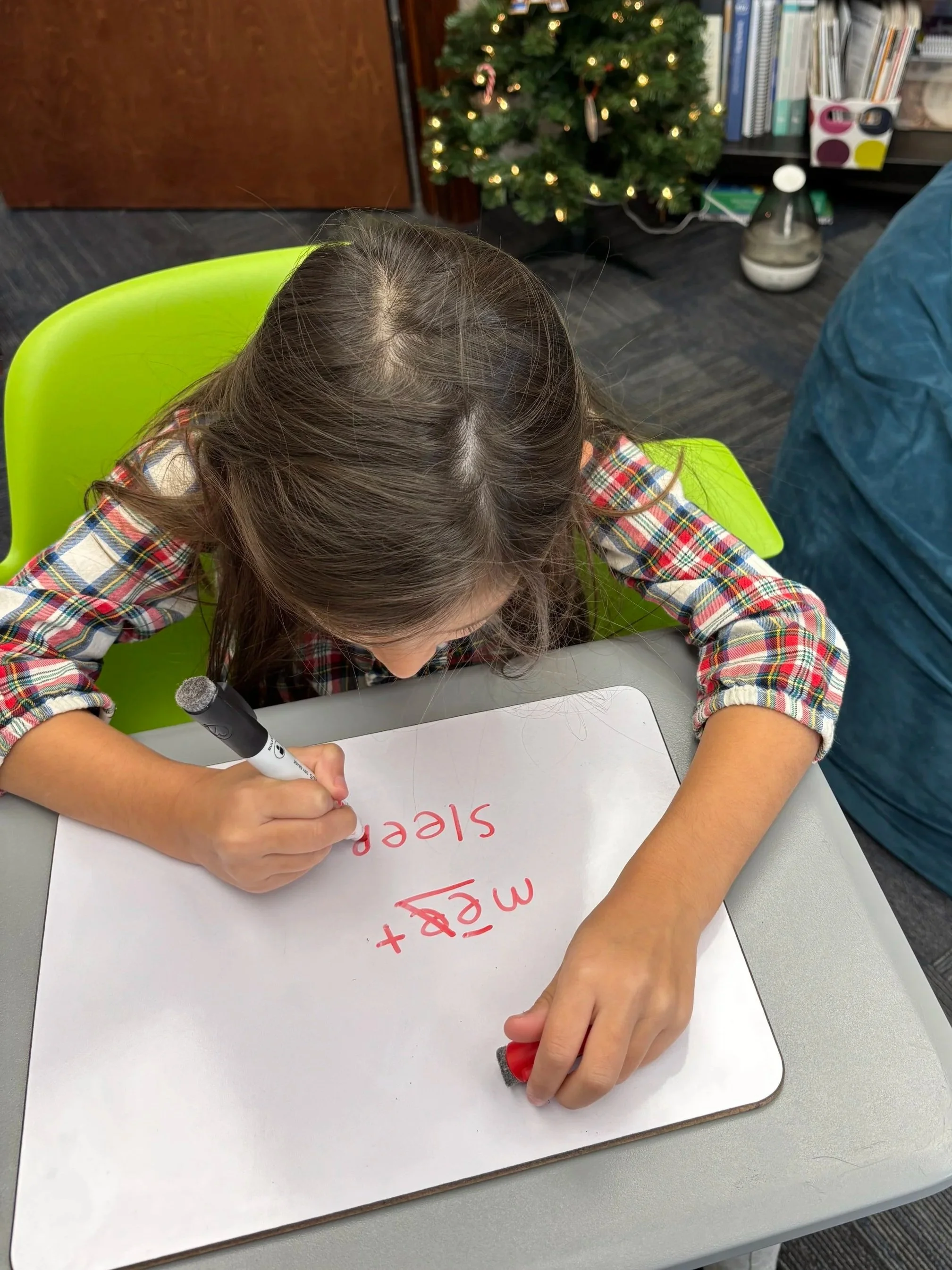 A young girl writing on a whiteboard with a red marker, the words 'ME+T+E+S P' are visible, sitting at a desk with a green chair, a Christmas tree and office shelves in the background.