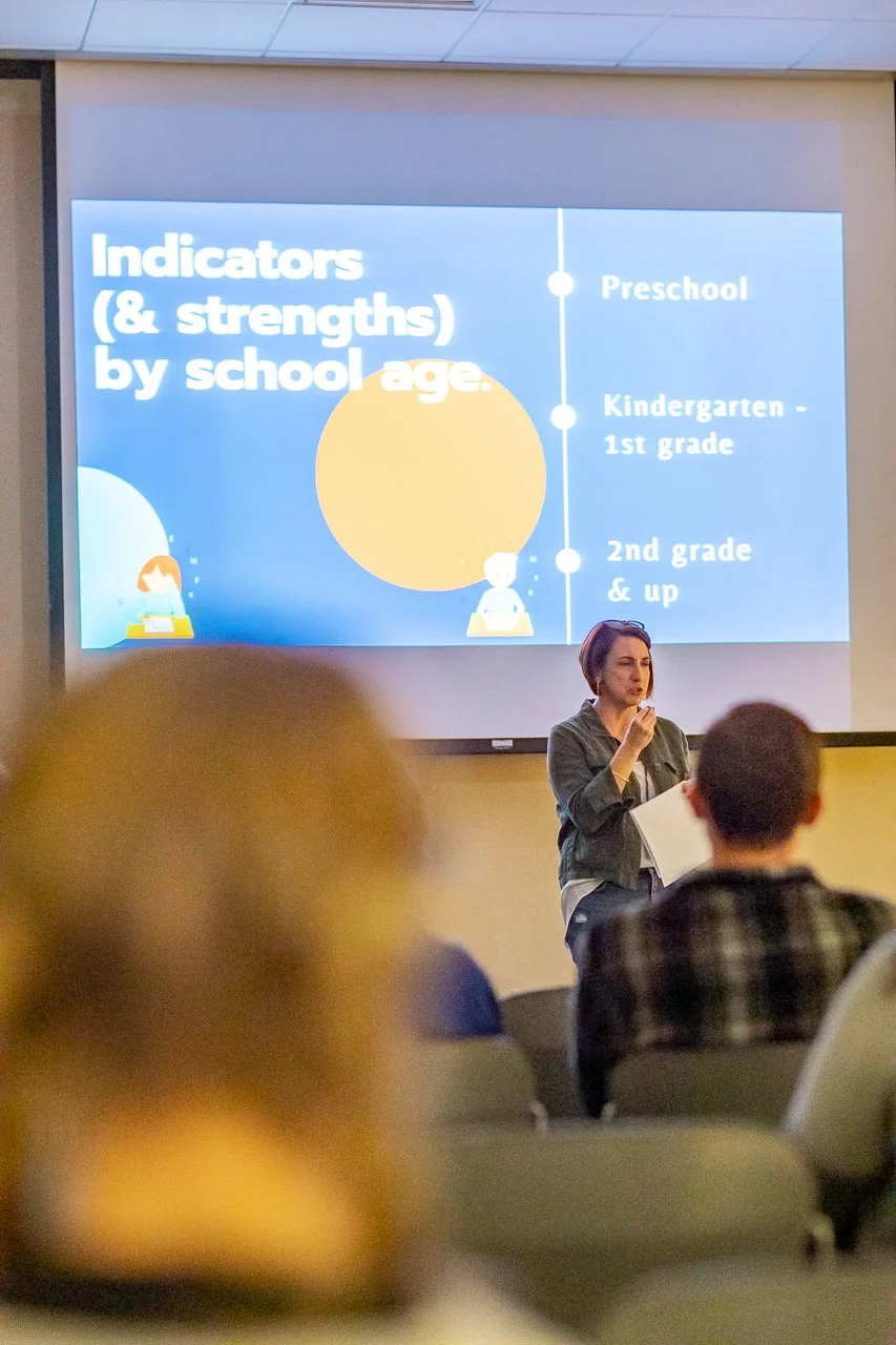 A woman giving a presentation about indicators and strengths by school age, with a slide showing categories: preschool, kindergarten to 1st grade, and 2nd grade and up, in a classroom or meeting room.