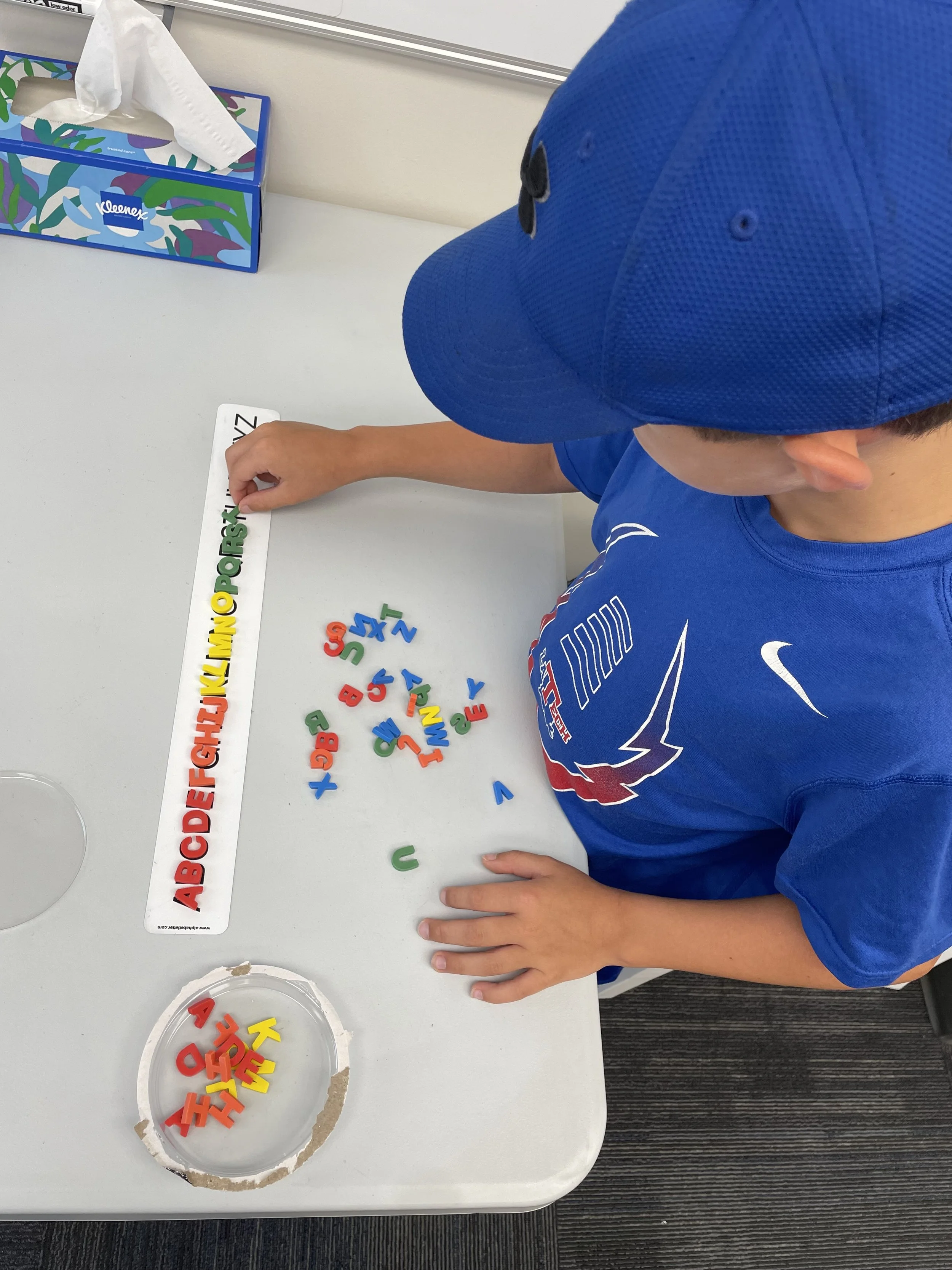A young boy wearing a blue cap and a blue t-shirt is sitting at a white table, arranging colorful foam letters and numbers beside a strip chart with the alphabet written on it. There is a small round container with additional foam letters on the tabl