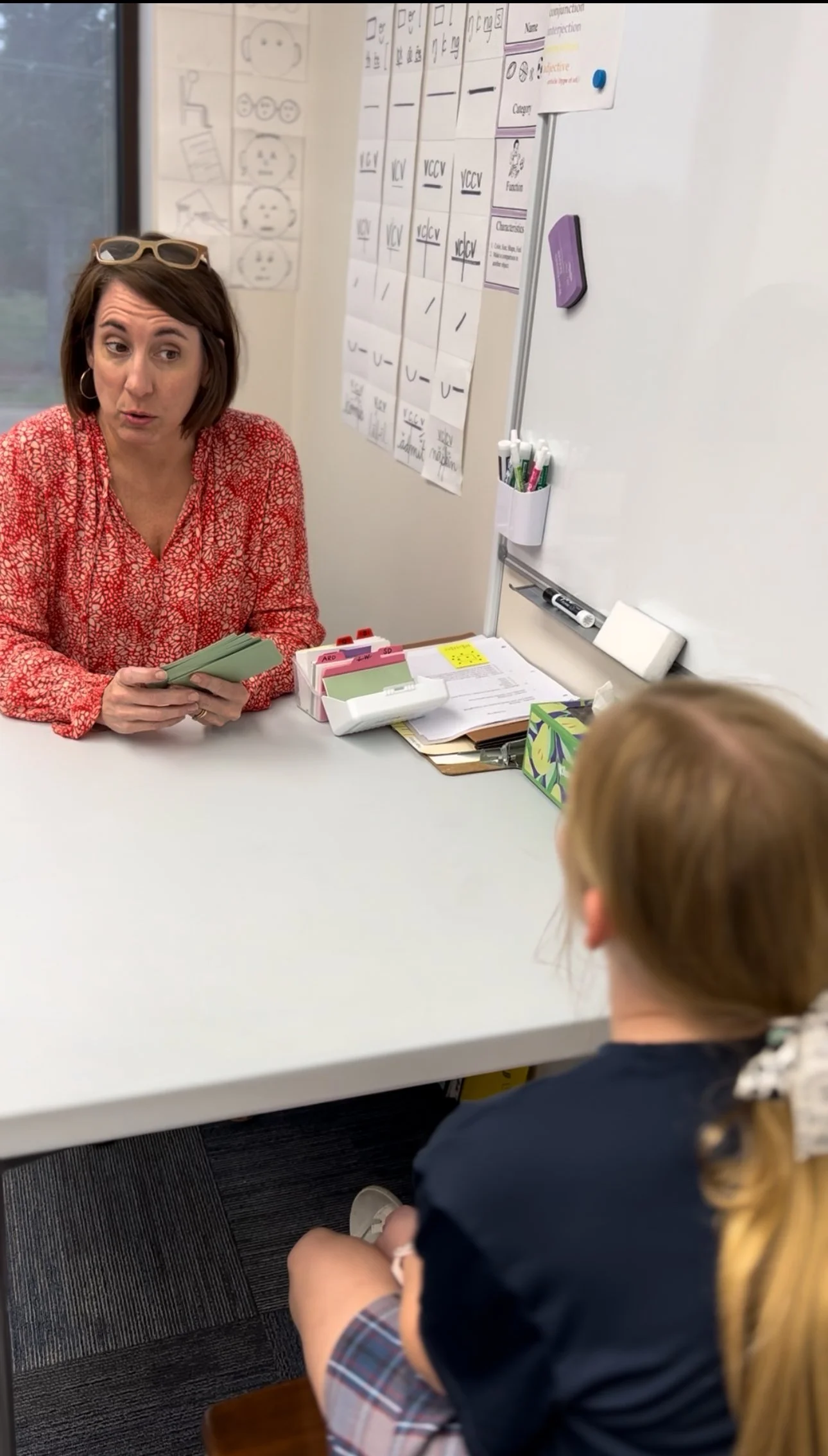 A woman with glasses on her head and hoop earrings is sitting across from a young girl at a white table. The woman is holding a hand of playing cards and appears to be talking. The girl has blonde hair tied with a ribbon and is wearing a school unifo