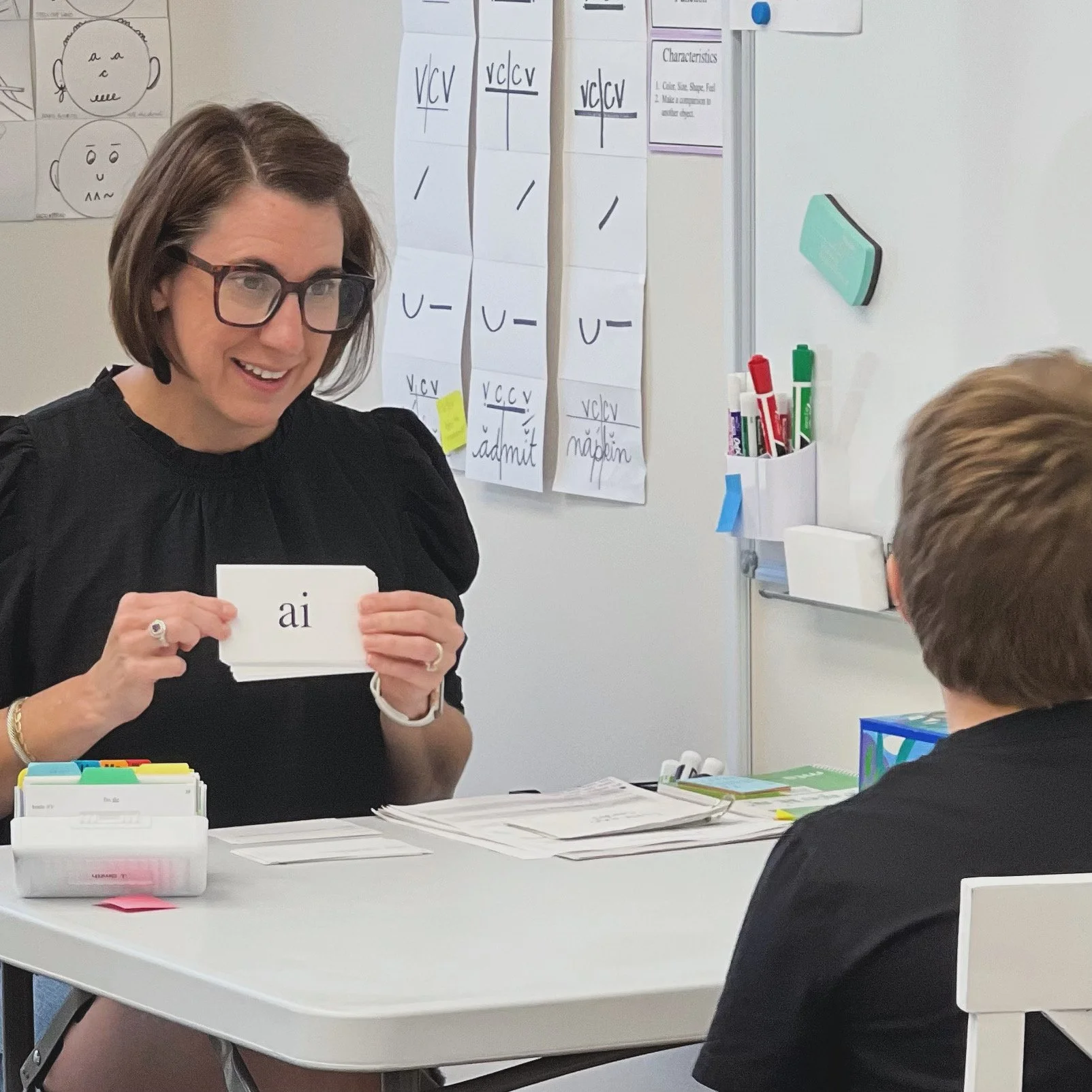 A woman in glasses and black shirt holding flashcards with the phonetic spelling 'ai' while sitting at a table with a young boy in a classroom. There are educational posters and supplies on the table and whiteboard in the background.