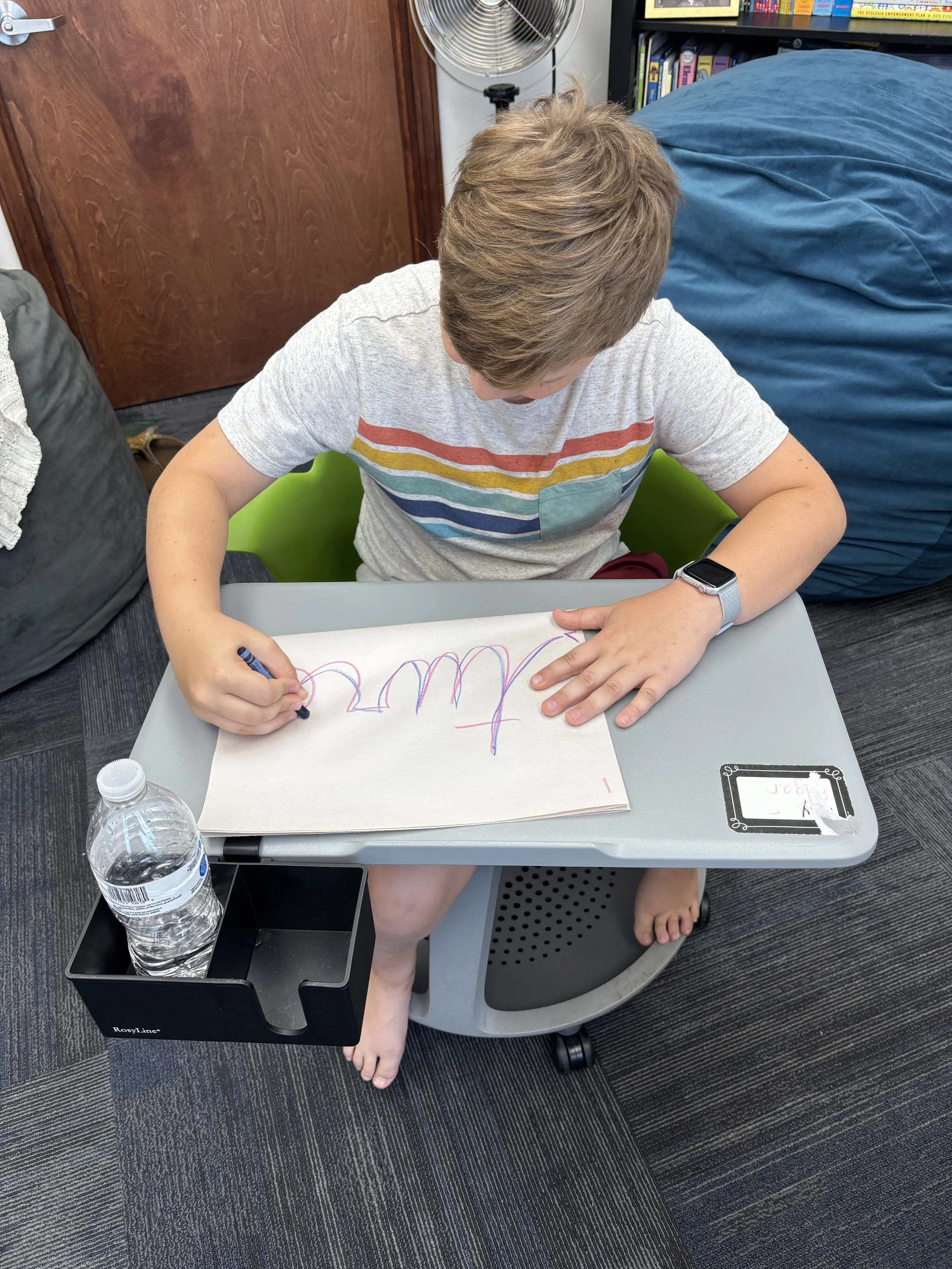 A young boy with light brown hair, wearing a gray t-shirt with colorful horizontal stripes and a watch on his left wrist, is sitting at a small gray desk. He is drawing on a large white sheet of paper with multi-colored lines. The desk has a built-in