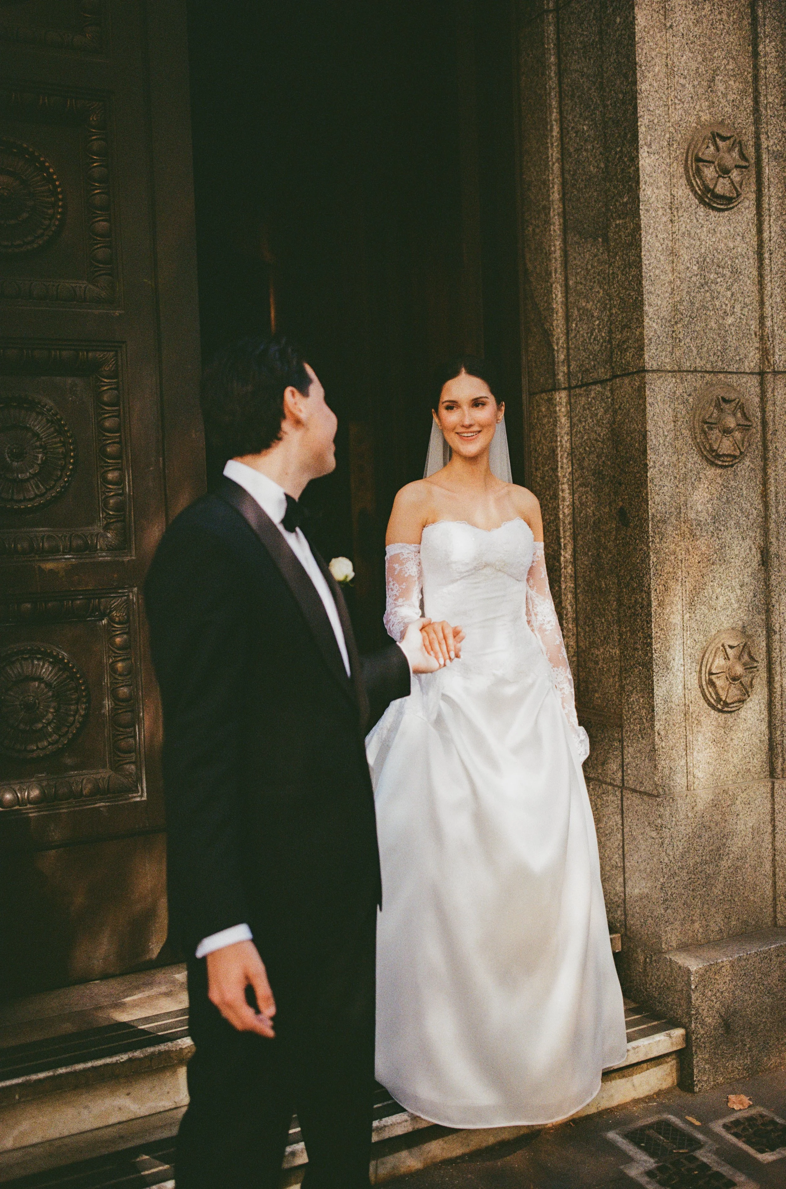 A woman in a strapless white wedding dress with lace sleeves is holding a man's hand. The man is wearing a black tuxedo with a white shirt and black bow tie. They are standing outside a building with decorative stone and metal features.