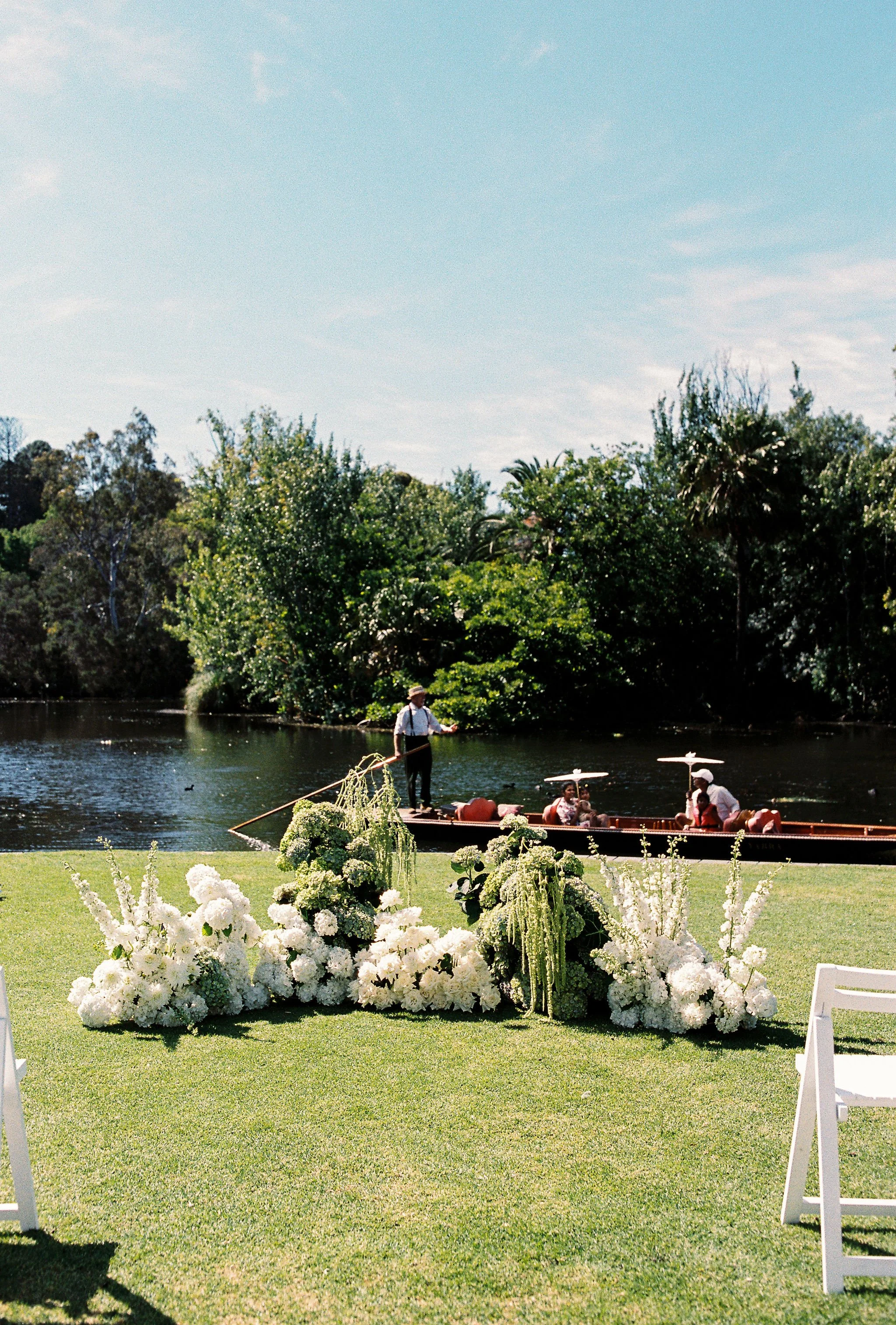 A boat with passengers floating on a river with trees in the background and a grassy area decorated with white flowers in the foreground.