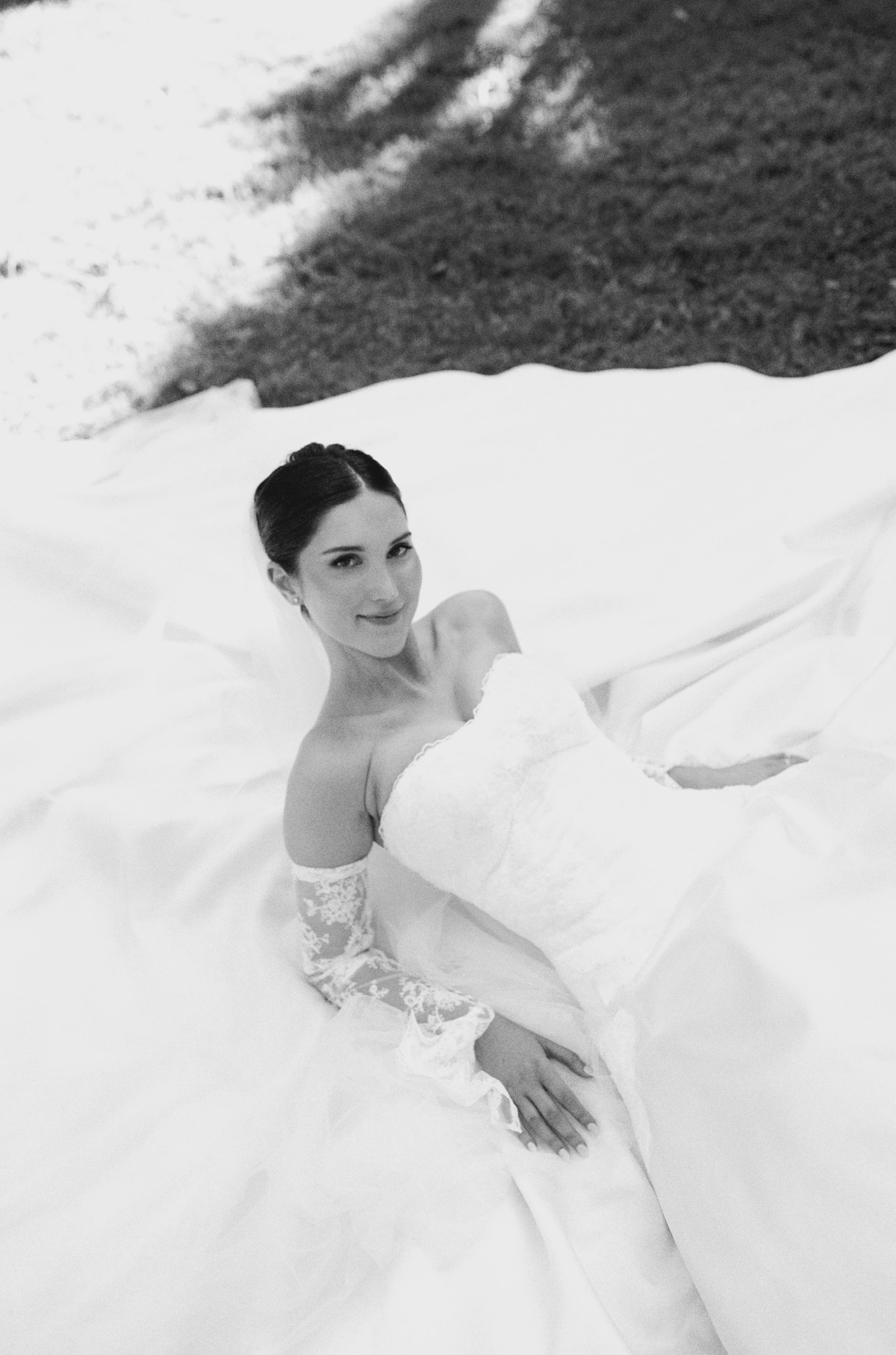 A woman in a wedding dress sitting on a white bed, smiling, holding her leg, with dark hair, lace sleeves, and a strapless bodice, in a bedroom with a dark wall.