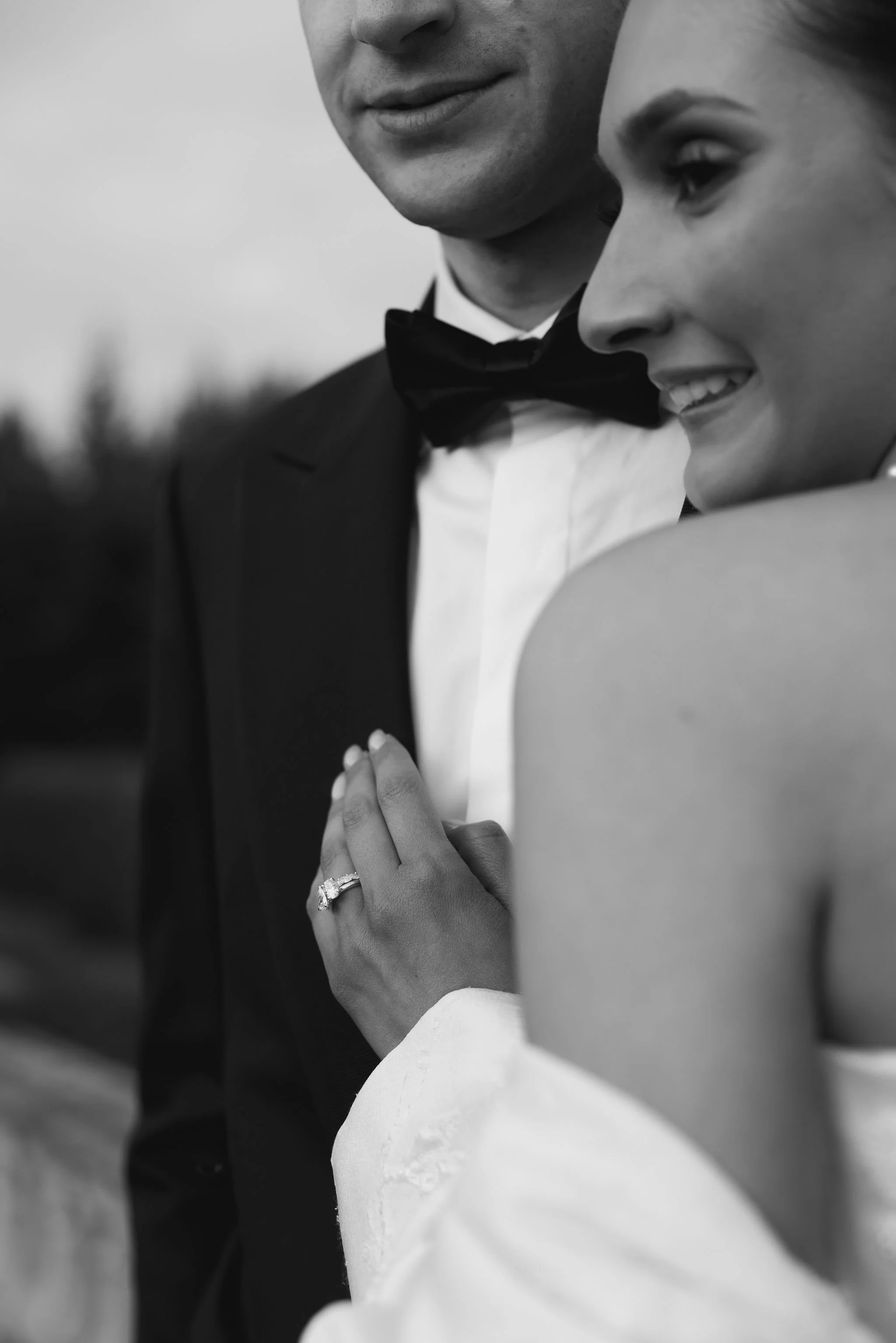 Close-up black and white photo of a couple, where the woman is smiling and the man is only partially visible, both dressed formally, possibly at a wedding, with the woman wearing a ring on her finger.