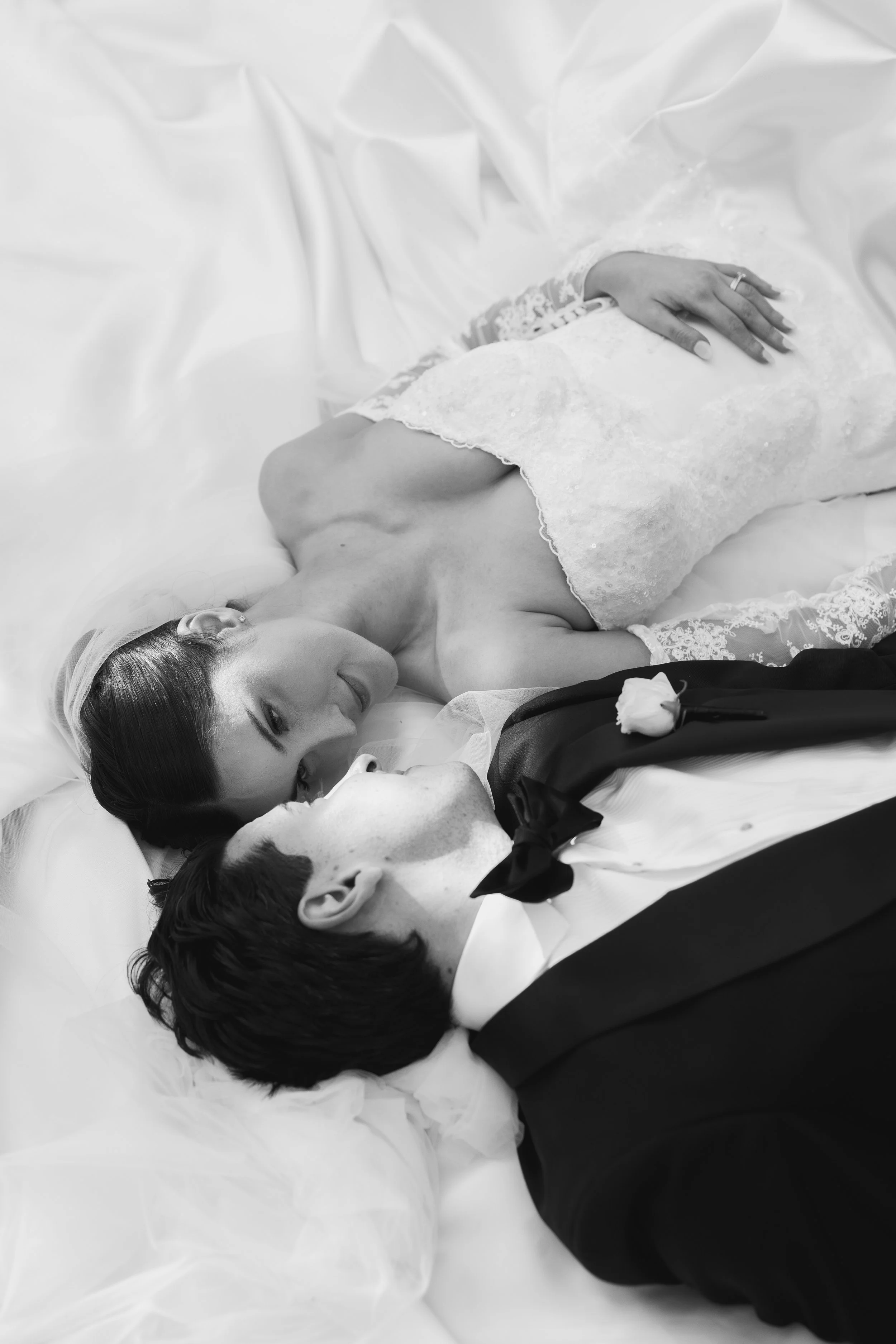 A black and white photo of a bride and groom lying on a bed, gazing at each other, with the bride wearing lace and the groom in a tuxedo. Their hands are close, and a single rose is on the bride's side.