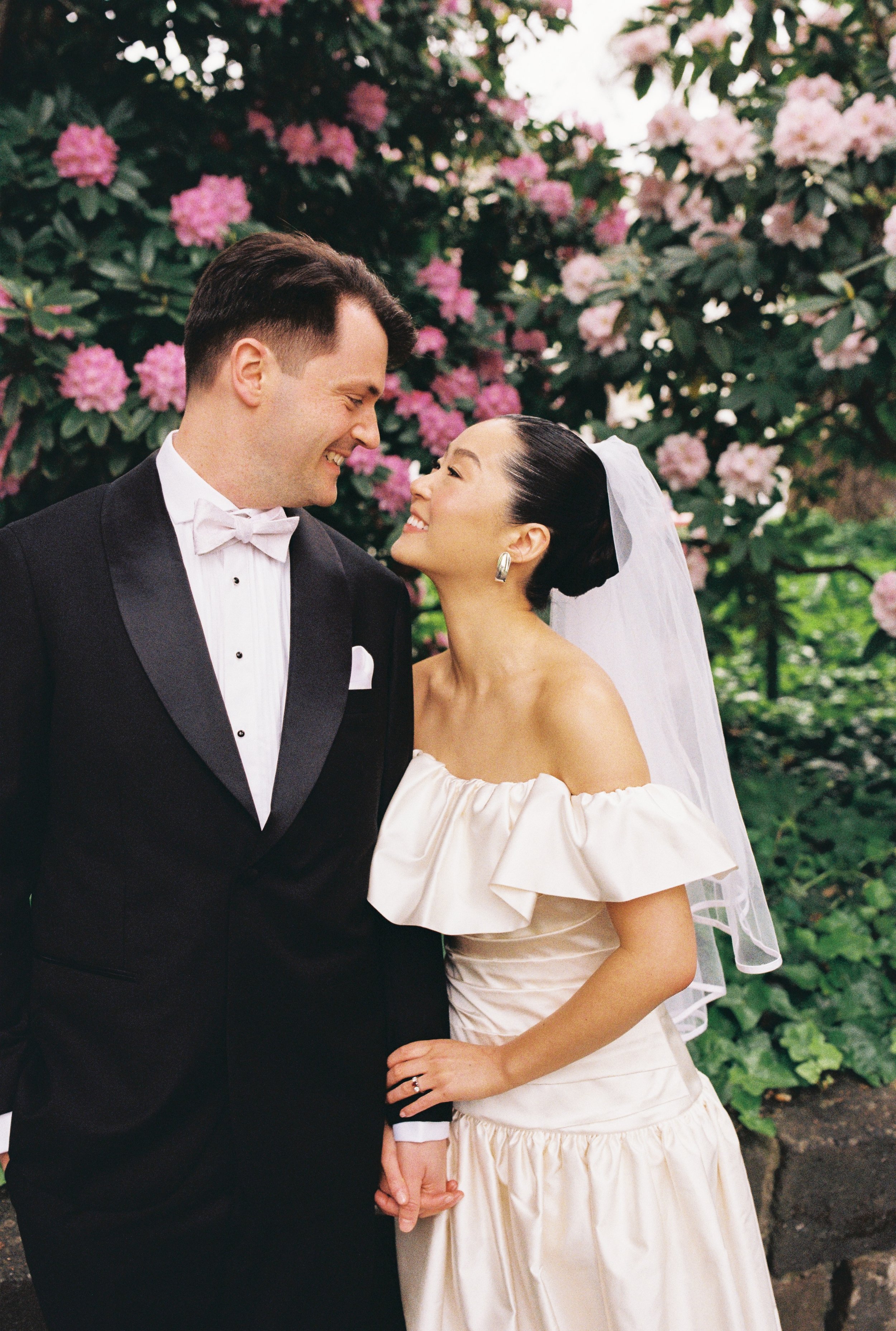 A smiling newlywed couple holding hands outdoors in front of pink and white flowering bushes, with the groom wearing a black tuxedo and the bride wearing a white off-shoulder wedding dress and veil.