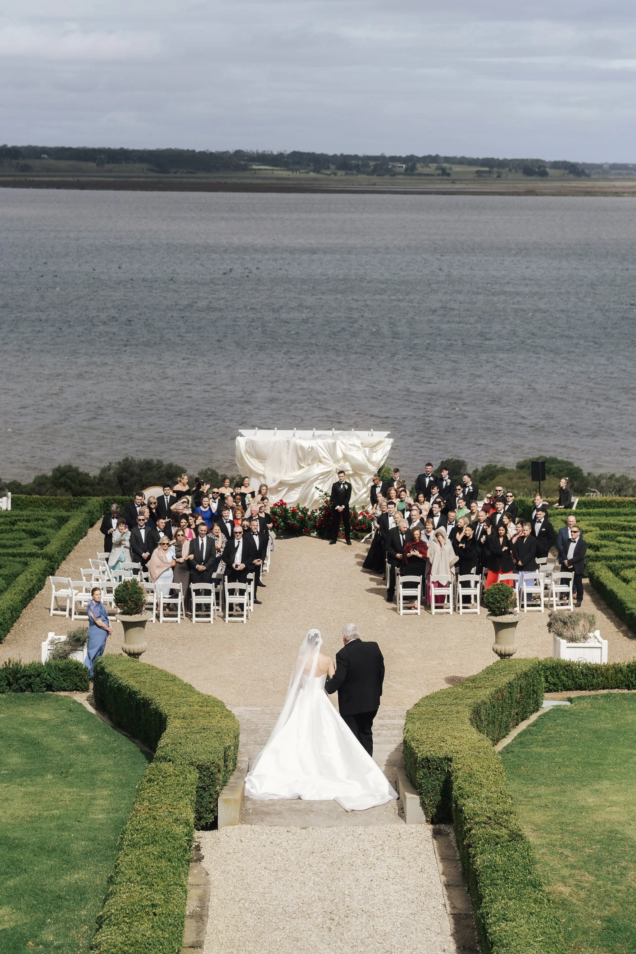A wedding ceremony outdoors with a bride and groom walking down the aisle, surrounded by guests and a scenic water view in the background.