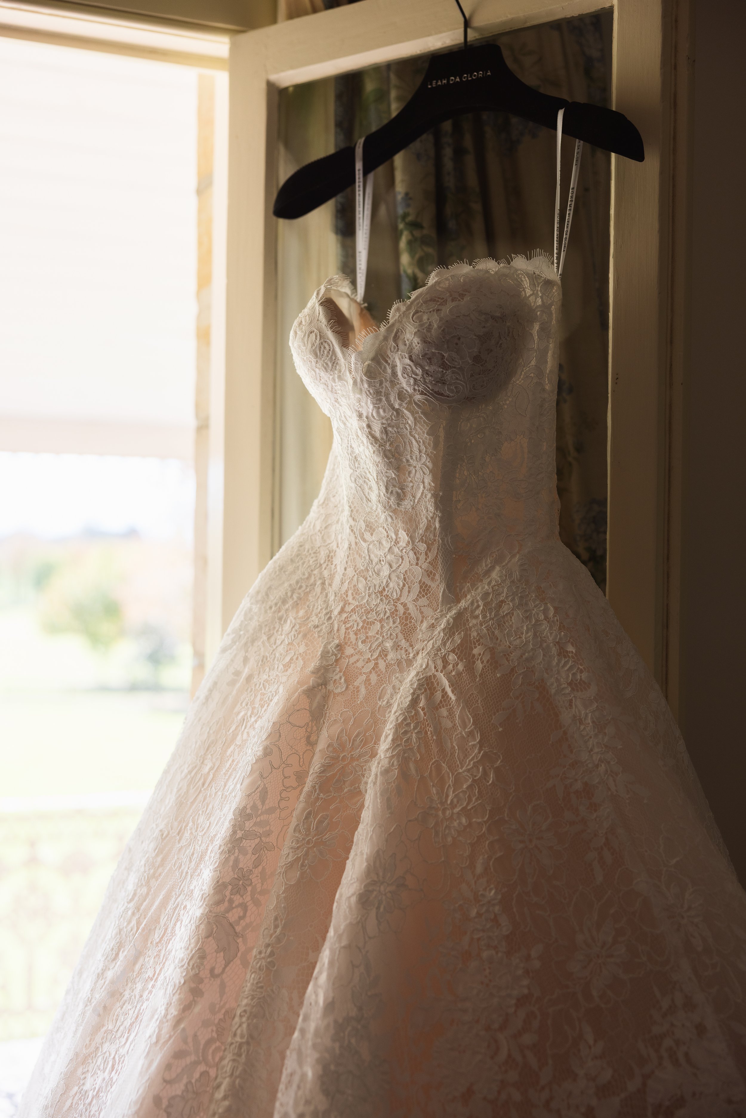 A wedding dress with lace detailing hanging on a black hanger inside a wooden frame, with sunlight streaming in from a window in the background.