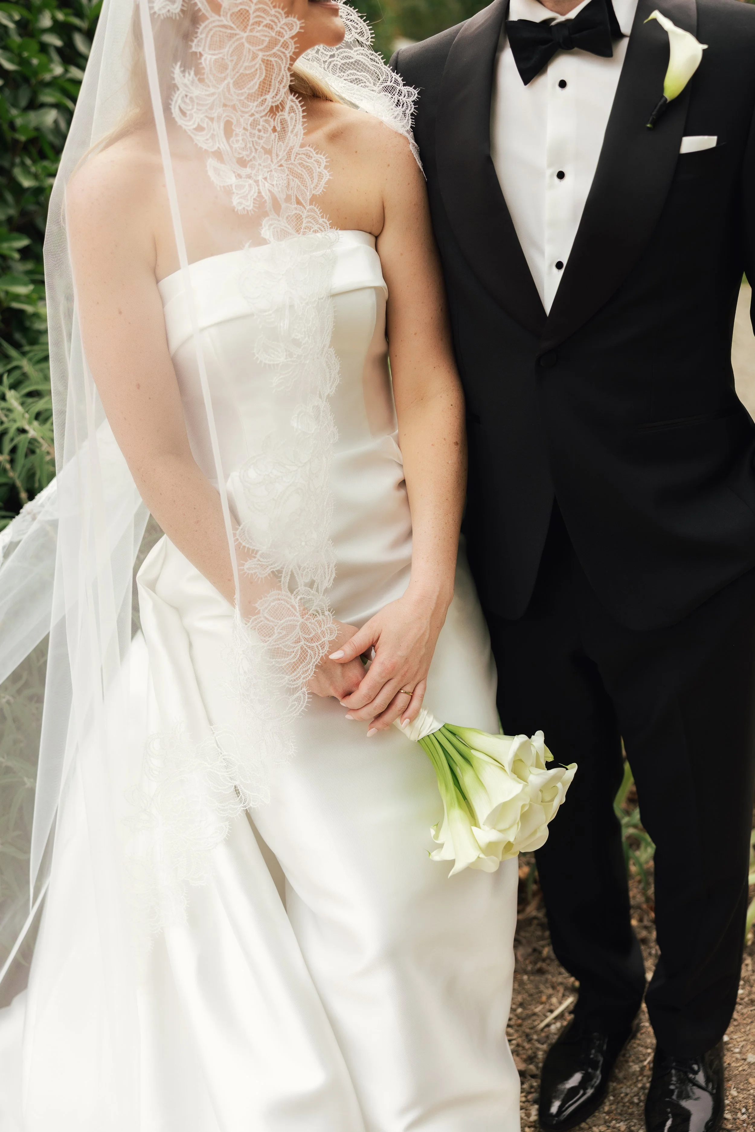 A bride and groom standing closely at their wedding, with the bride in a white gown holding a bouquet of white calla lilies, and the groom in a black tuxedo with a white boutonniere.