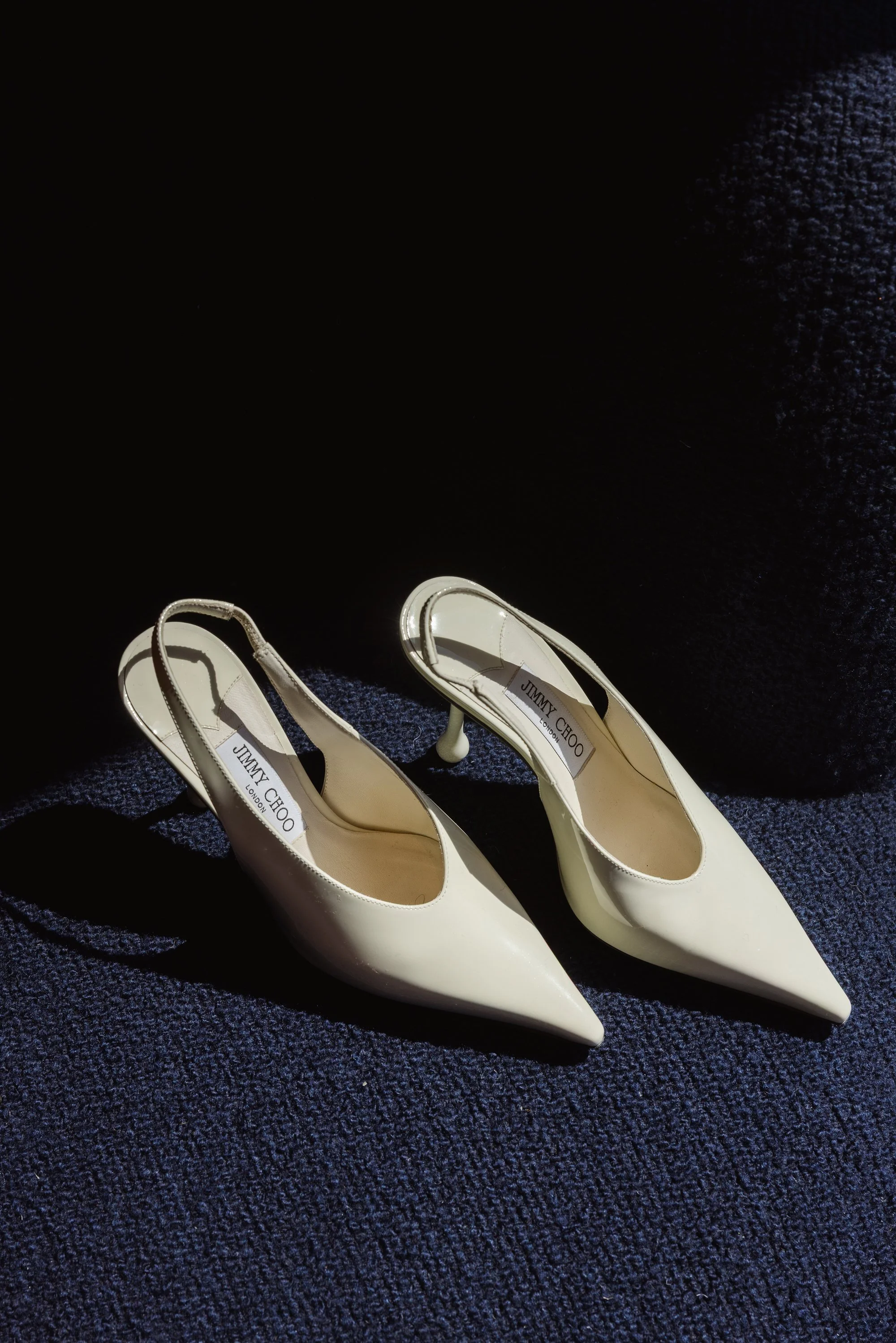 A pair of white Jimmy Choo high-heeled shoes with pointed toes on a dark textured surface.