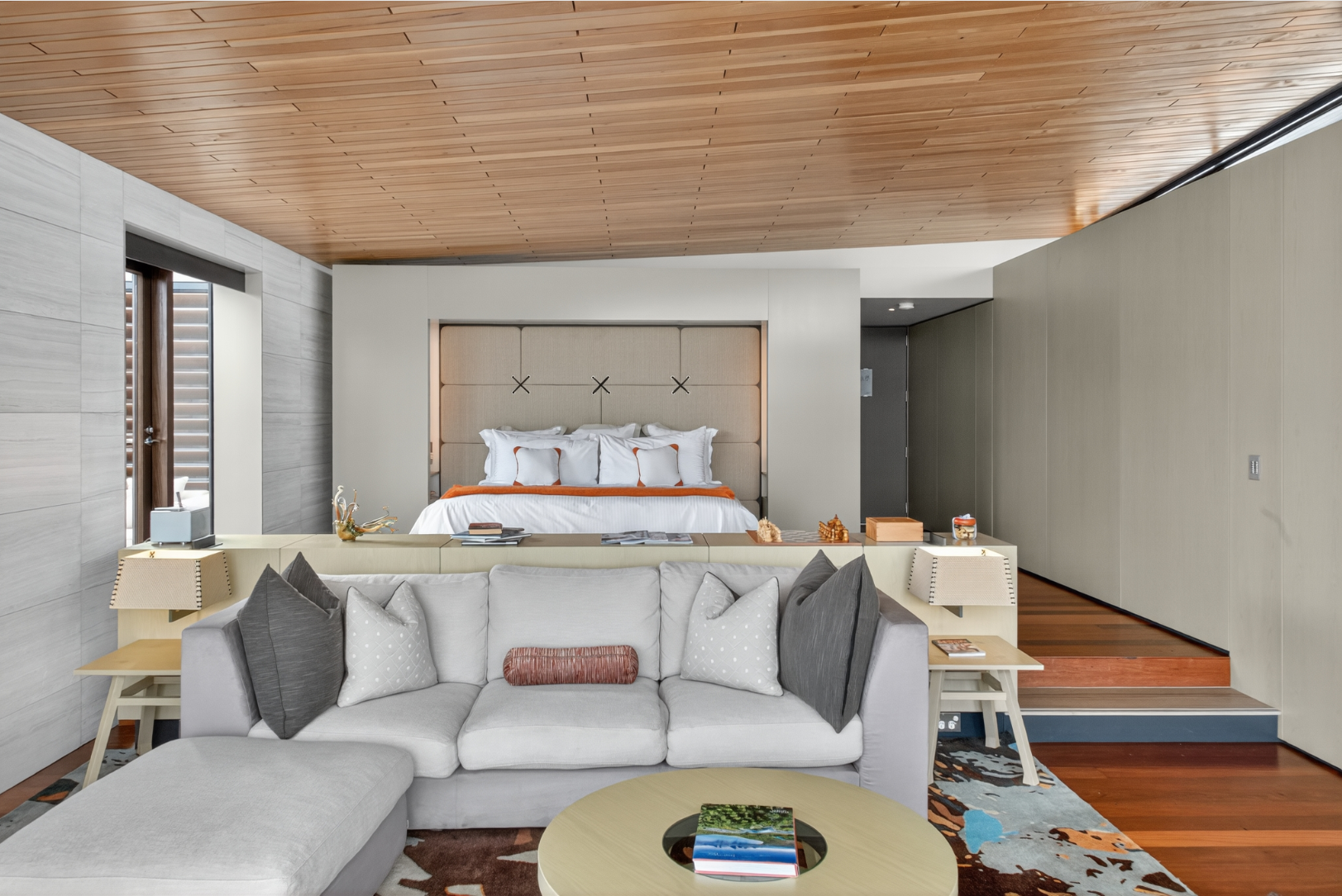Modern hotel suite featuring a cozy living room with a sectional sofa, decorative pillows, and a coffee table, overlooking a bedroom with a large bed, tufted headboard, and wooden ceiling.