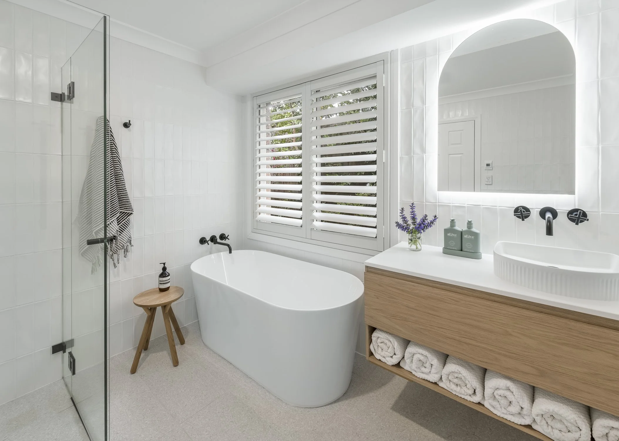Modern bathroom with white subway tiles, a freestanding bathtub, a window with white shutters, a wooden vanity with rolled towels underneath, a large mirror with backlighting, and a glass-enclosed shower area.