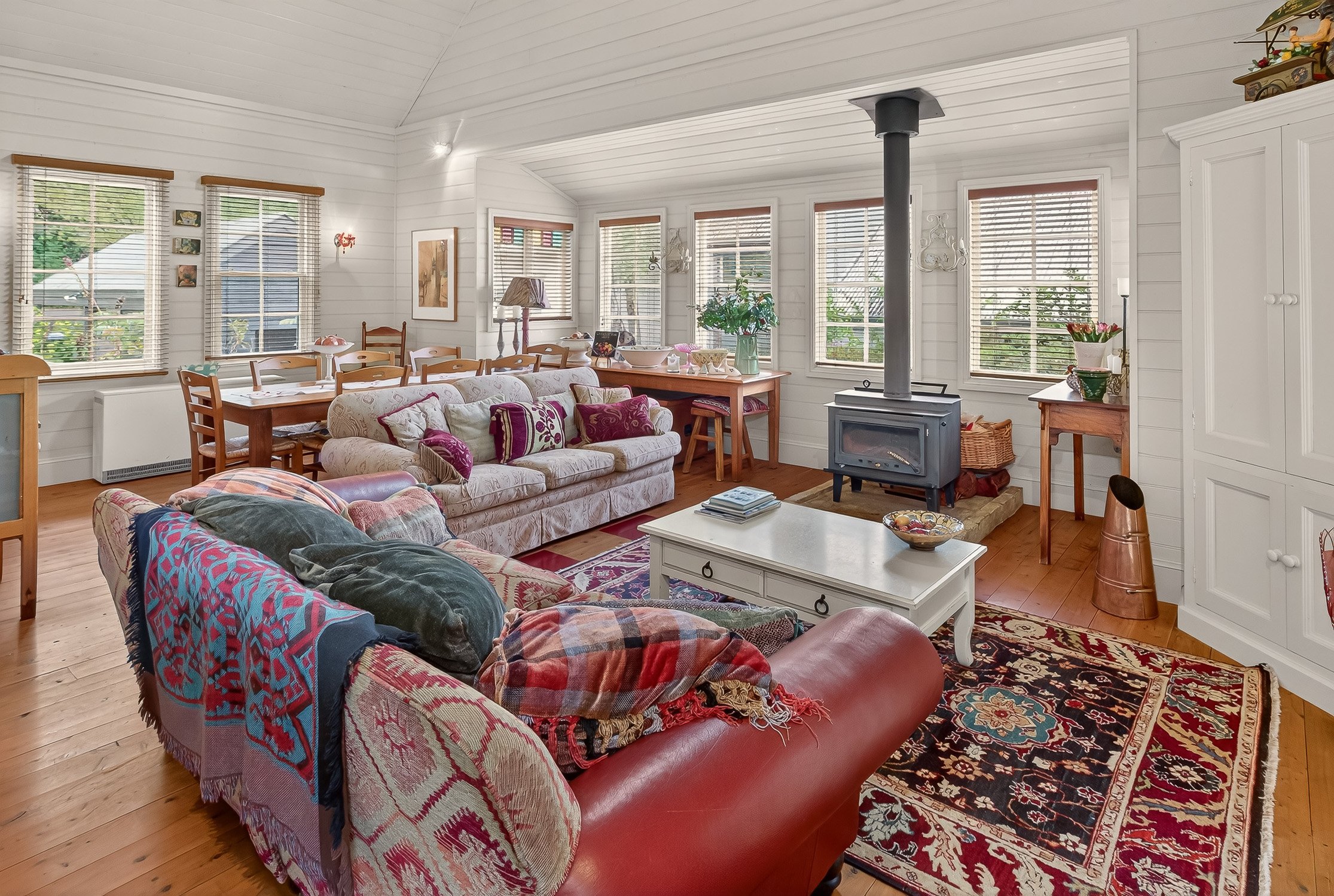 Interior of a bright living room with white wooden walls, multiple windows with blinds, a wood stove, a floral sofa with pillows, a red leather armchair with blankets, a white coffee table, and a patterned rug.