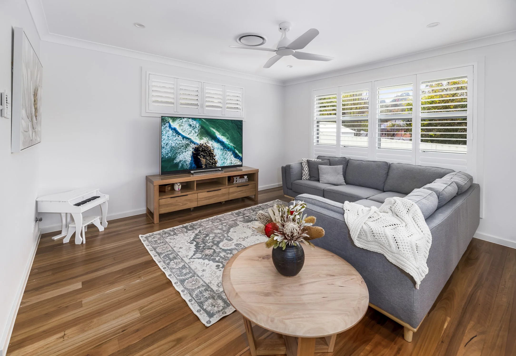 Living room with white walls, wooden floor, gray sectional sofa, round wooden coffee table with flower arrangement, large windows with white shutters, flat-screen TV on wooden stand, decorative rug, ceiling fan, and small white piano.