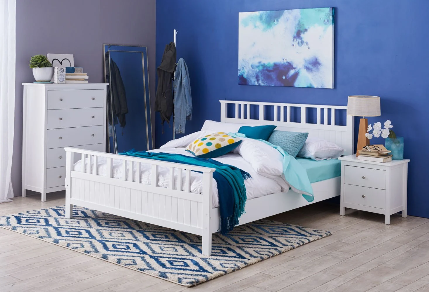 A bedroom with blue and white decor, including a white bed with pillows and a throw blanket, a white dresser, a white nightstand with a lamp, a large mirror, a clothing rack, a colorful abstract painting on the wall, and a patterned rug on light-colo