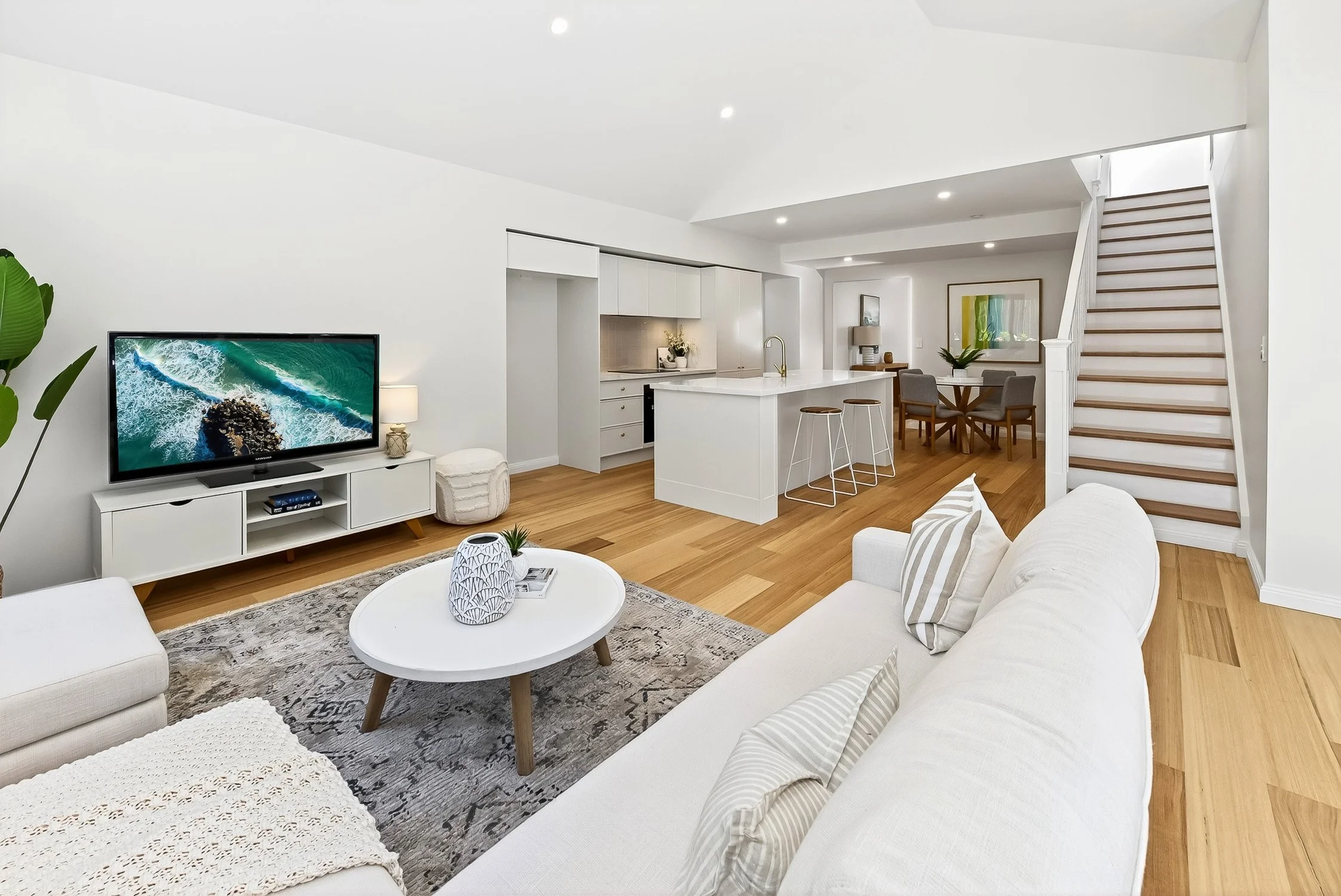 Open-concept living room with white sofa, coffee table, TV, and kitchen with island and dining area, featuring light wood floors, white walls, and modern decor.