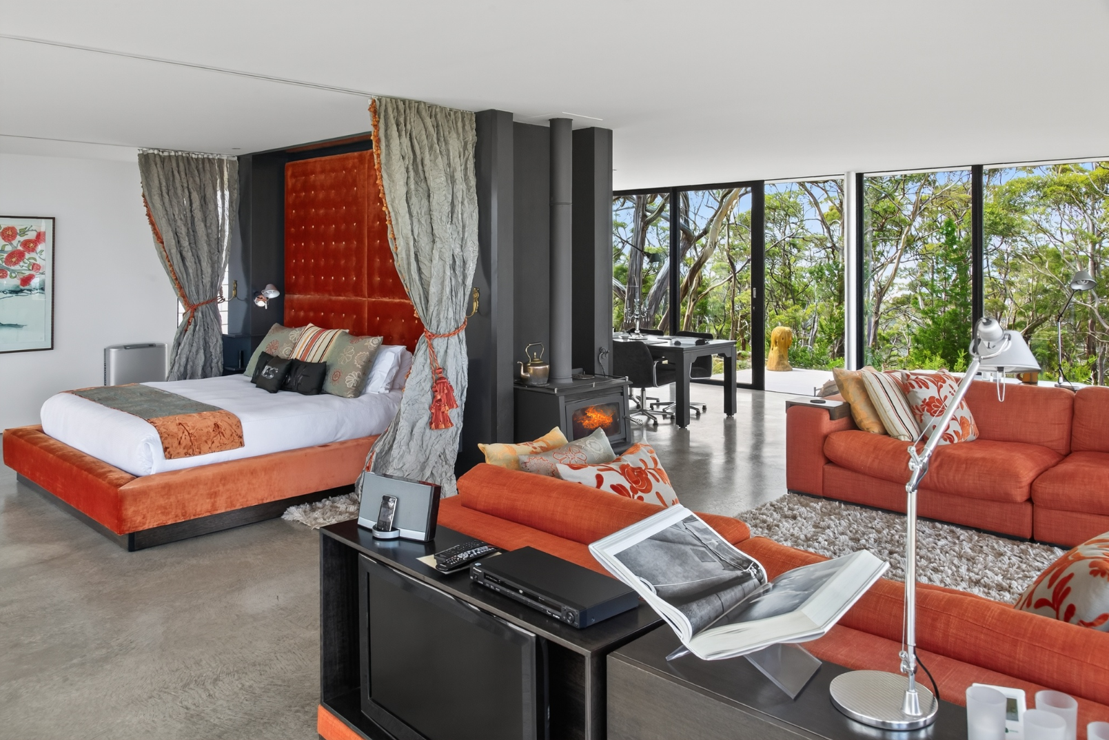 Modern bedroom with a large bed, orange and patterned cushions, and a red velvet headboard covered with fancy curtains. The room opens to a bright outdoor area with trees. There is a black fireplace, a black desk with a chair, and orange sofas with p