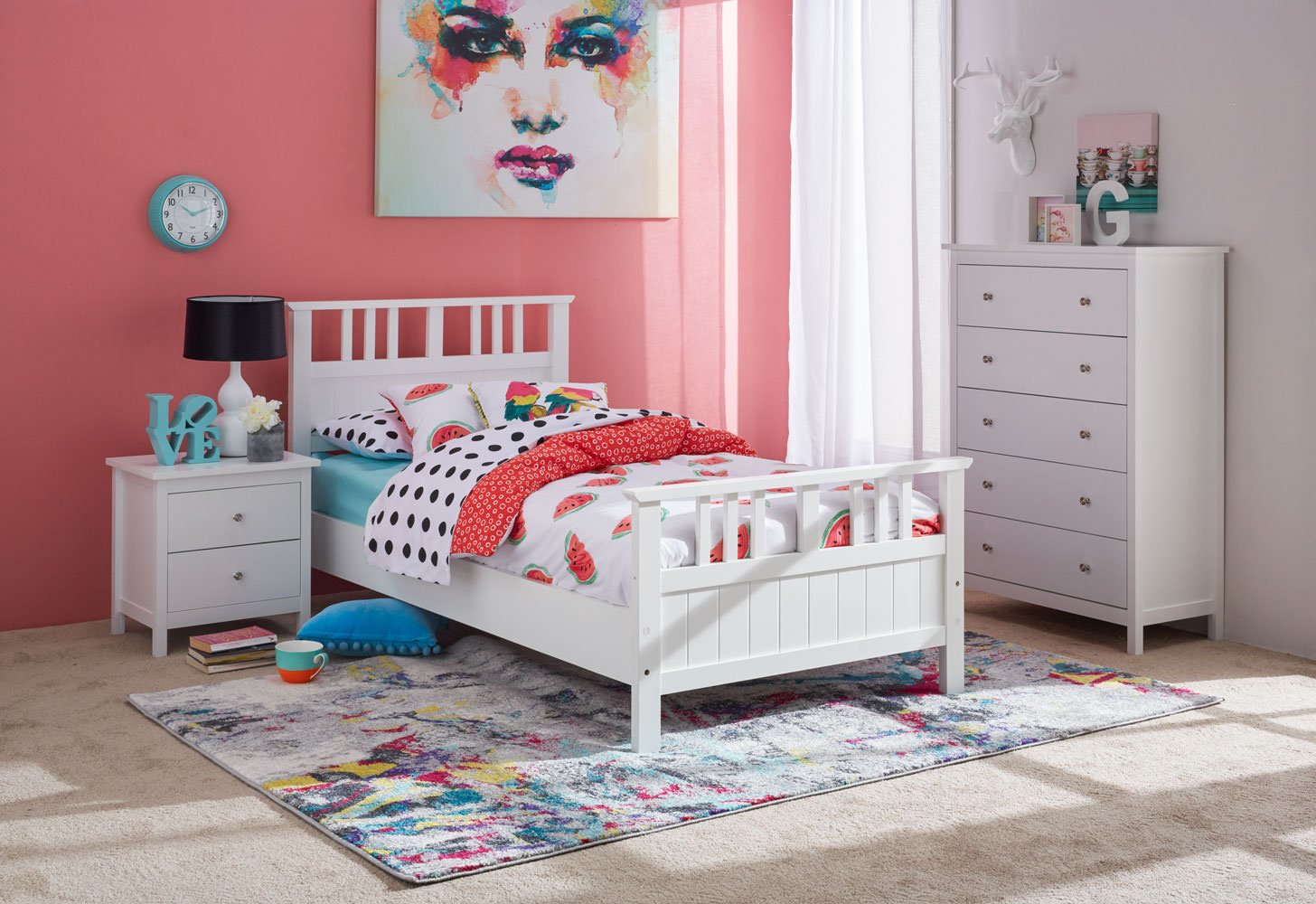 A colorful bedroom with a pink accent wall, featuring a white bed with a watermelon-themed bedspread, a nightstand with a blue decorative 'LOVE' sign, a black lamp, a round wall clock, a large abstract portrait above the bed, and a white dresser with