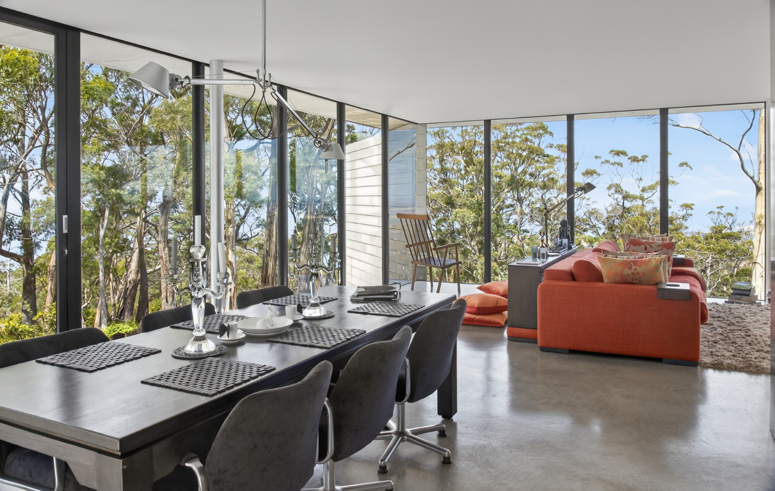 Modern dining and living room with large windows showing trees, a dining table set with placemats, candlesticks, and dishes, and a coral-colored sofa with pillows, a wooden chair, a shaggy rug, and a desk with a lamp and books.
