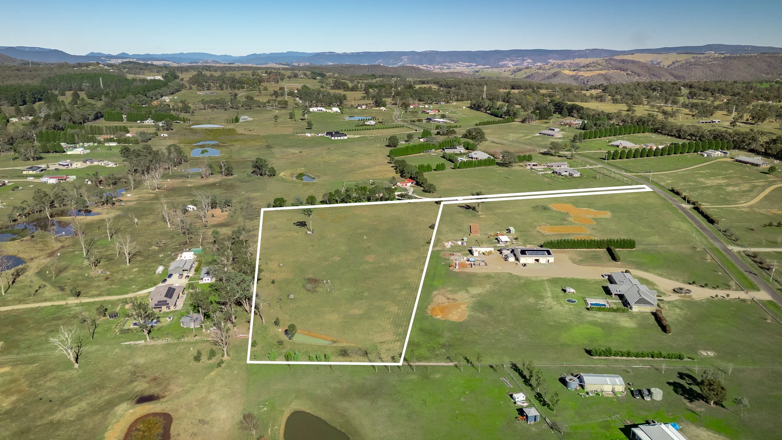 Drone Aerial view of a rural landscape in Hartley and Lithgow with green fields, small ponds, scattered houses, trees, and mountains in the background.