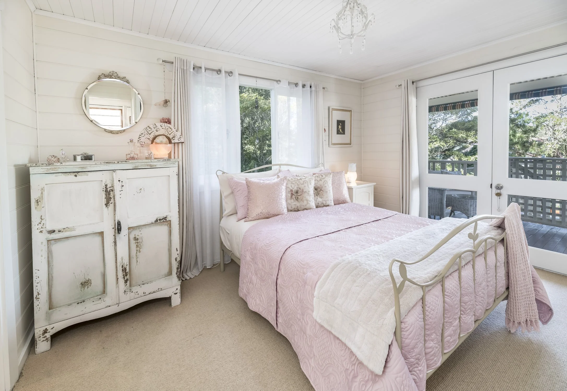 A bedroom with a white bed, pink quilt, and multiple pink and floral pillows. There are large windows with white curtains and a sliding glass door leading to a balcony with outdoor furniture. A distressed white cabinet is on the left side of the bed, and a small white nightstand with a lamp is on the right. The ceiling is white with a small chandelier.