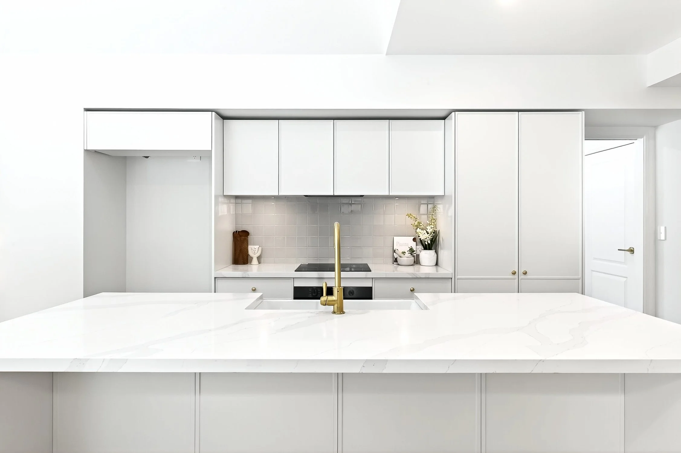 Modern white kitchen with marble island, gold faucet, white cabinets, and backsplash, decorated with plants and small accessories.