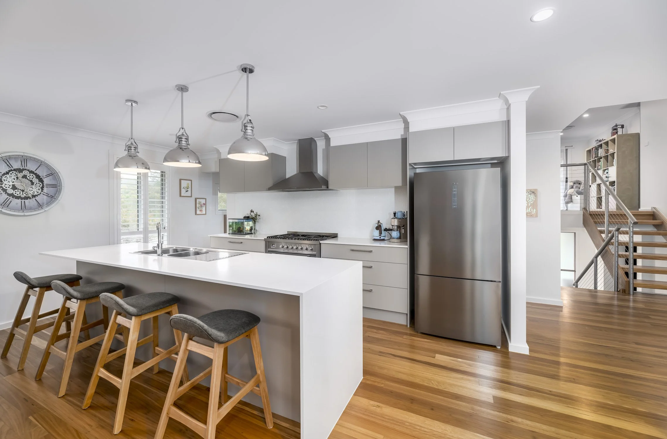 Modern kitchen with white countertops, gray cabinets, stainless steel refrigerator, and pendant lighting. Wooden bar stools with gray cushions are at the kitchen island. There is a clock on the wall and a fish tank on the counter.